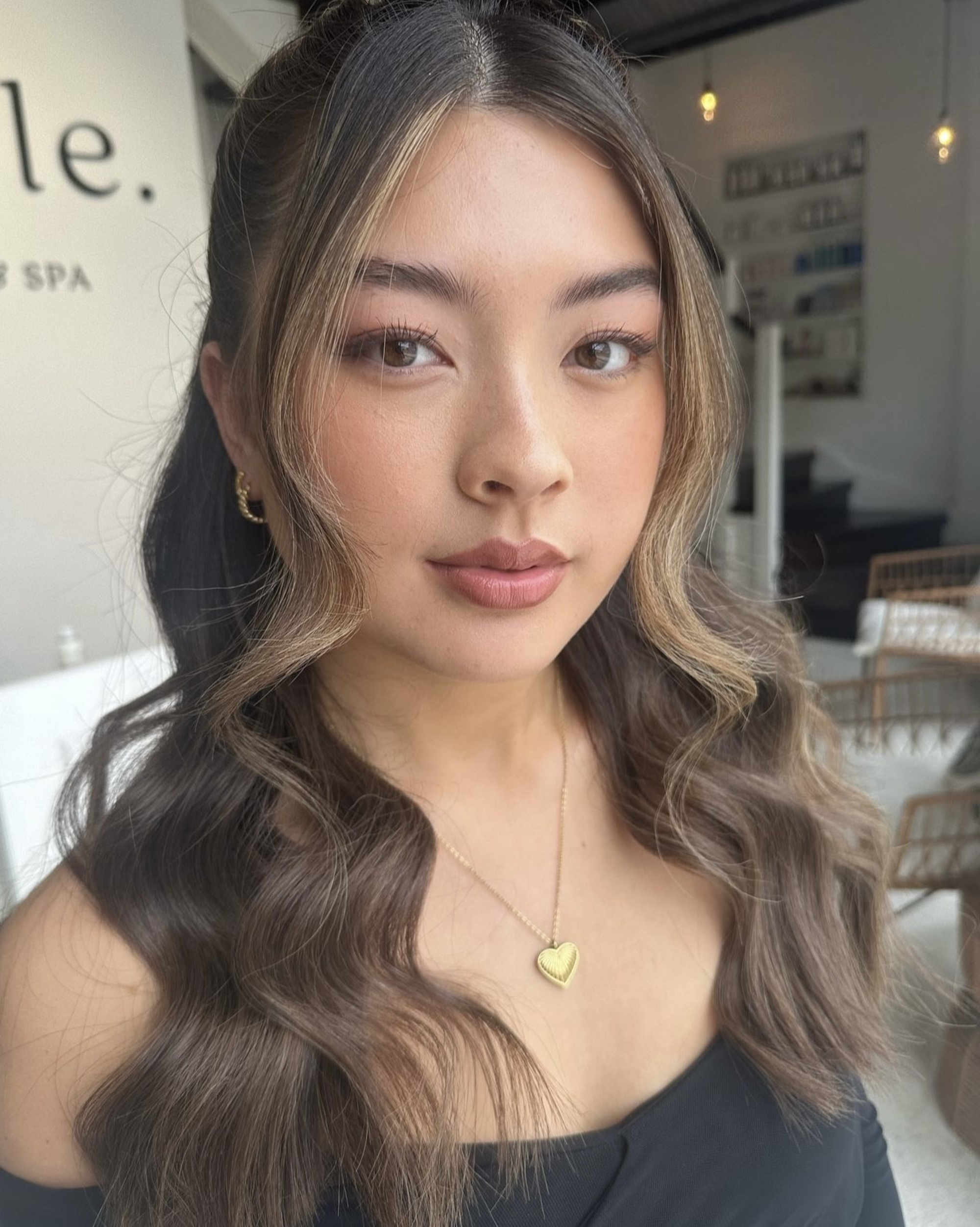 Close-up portrait of a woman with soft glam makeup and loose waves wearing a gold heart necklace in a bright studio.