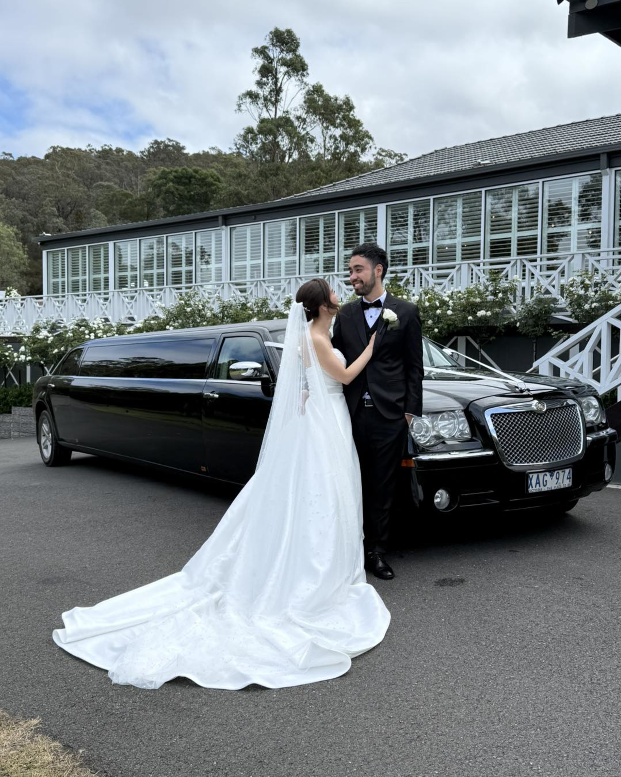 Bride and groom pose in front of a black stretch limousine outside a modern white wedding venue.