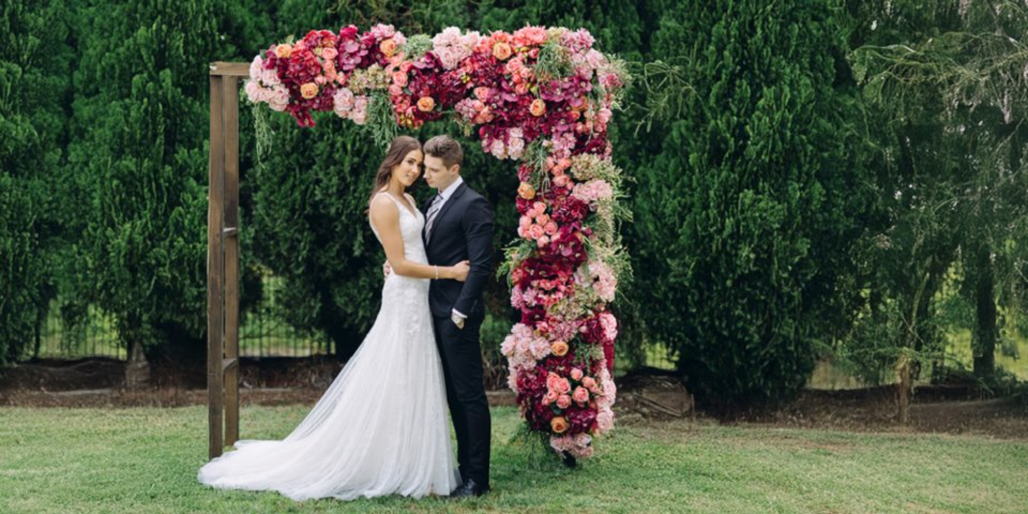 Bride and groom embrace beneath a lush pink floral ceremony arch in a green garden setting.