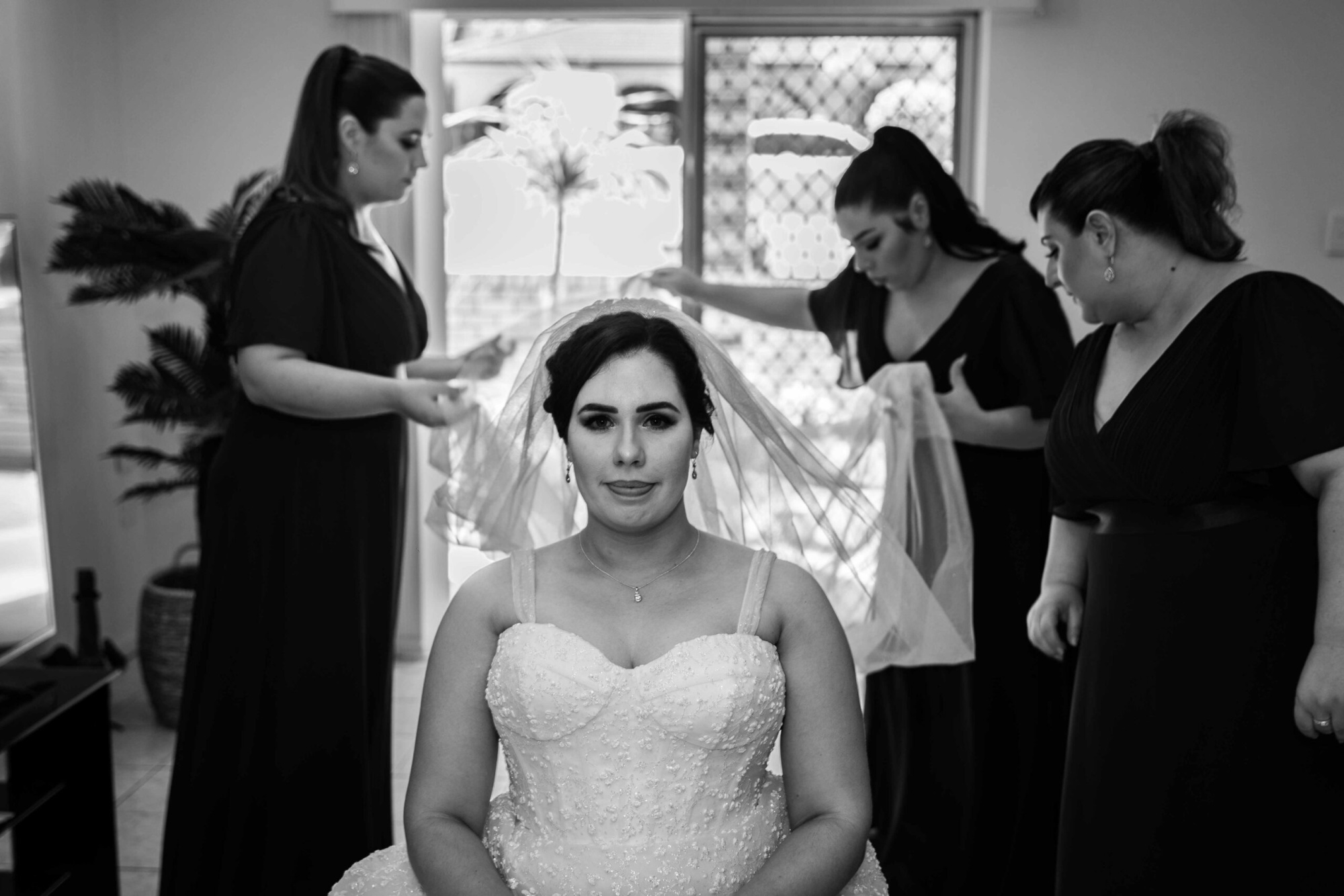 Bride sits in her gown while bridesmaids adjust her veil in a bright indoor setting.