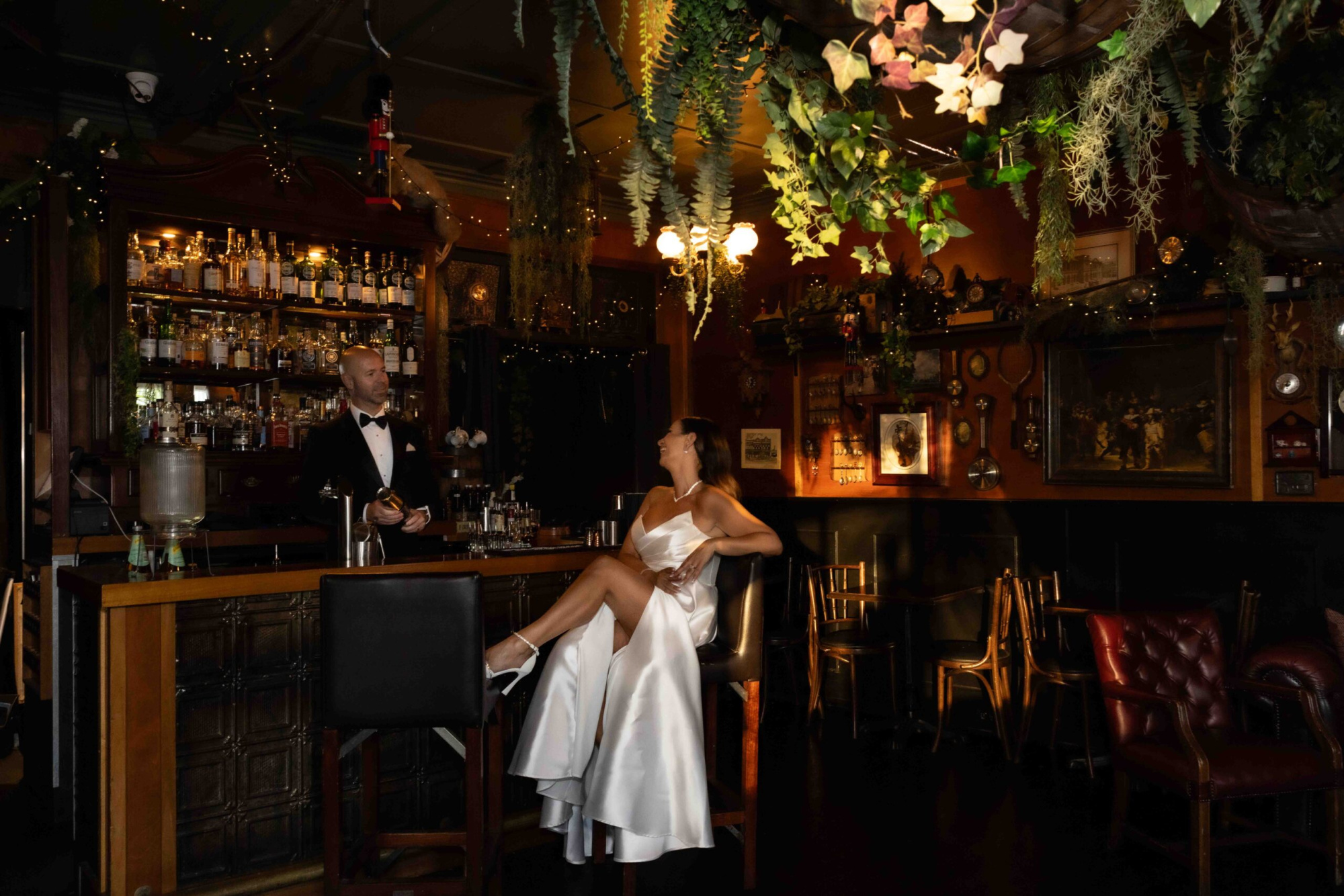 Bride in satin gown lounges at a moody cocktail bar while groom in tuxedo prepares a drink beneath hanging greenery.