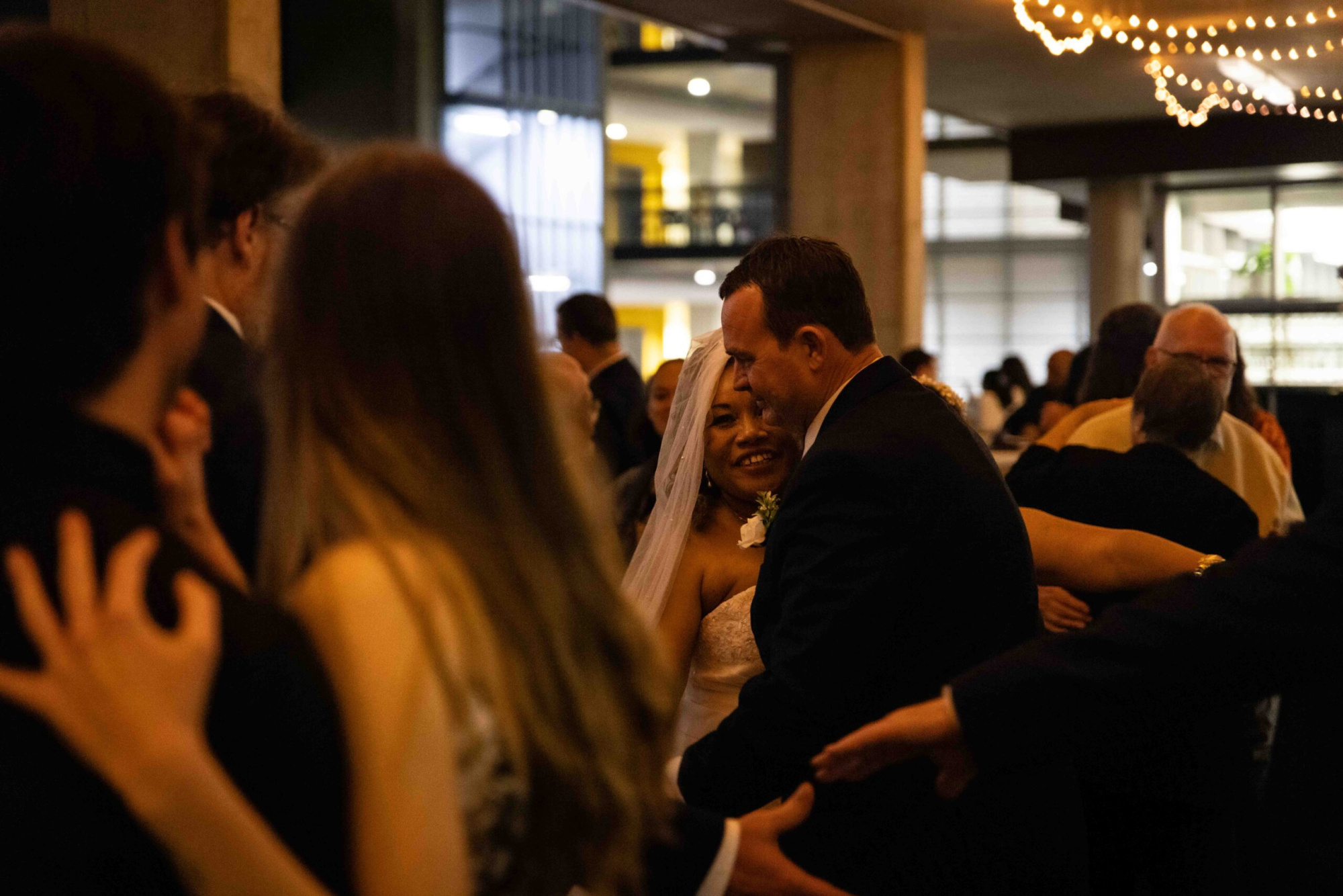 Bride and groom share a joyful dance surrounded by guests under warm string lights at an indoor wedding reception.