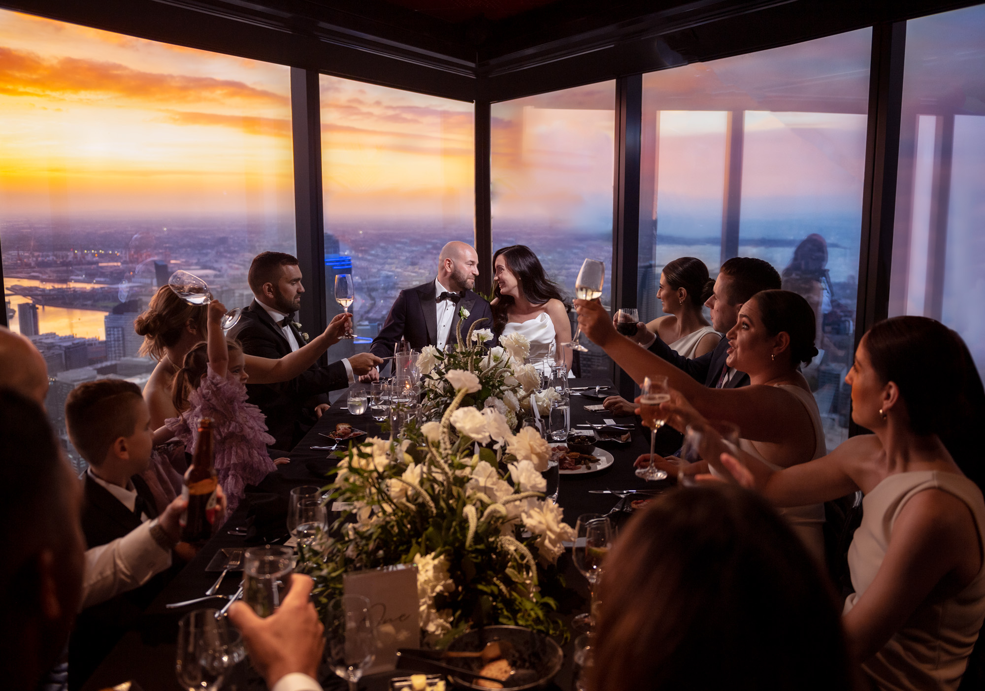 Wedding couple and guests share a sunset toast at an elegant city-view dinner table with floral centerpiece.
