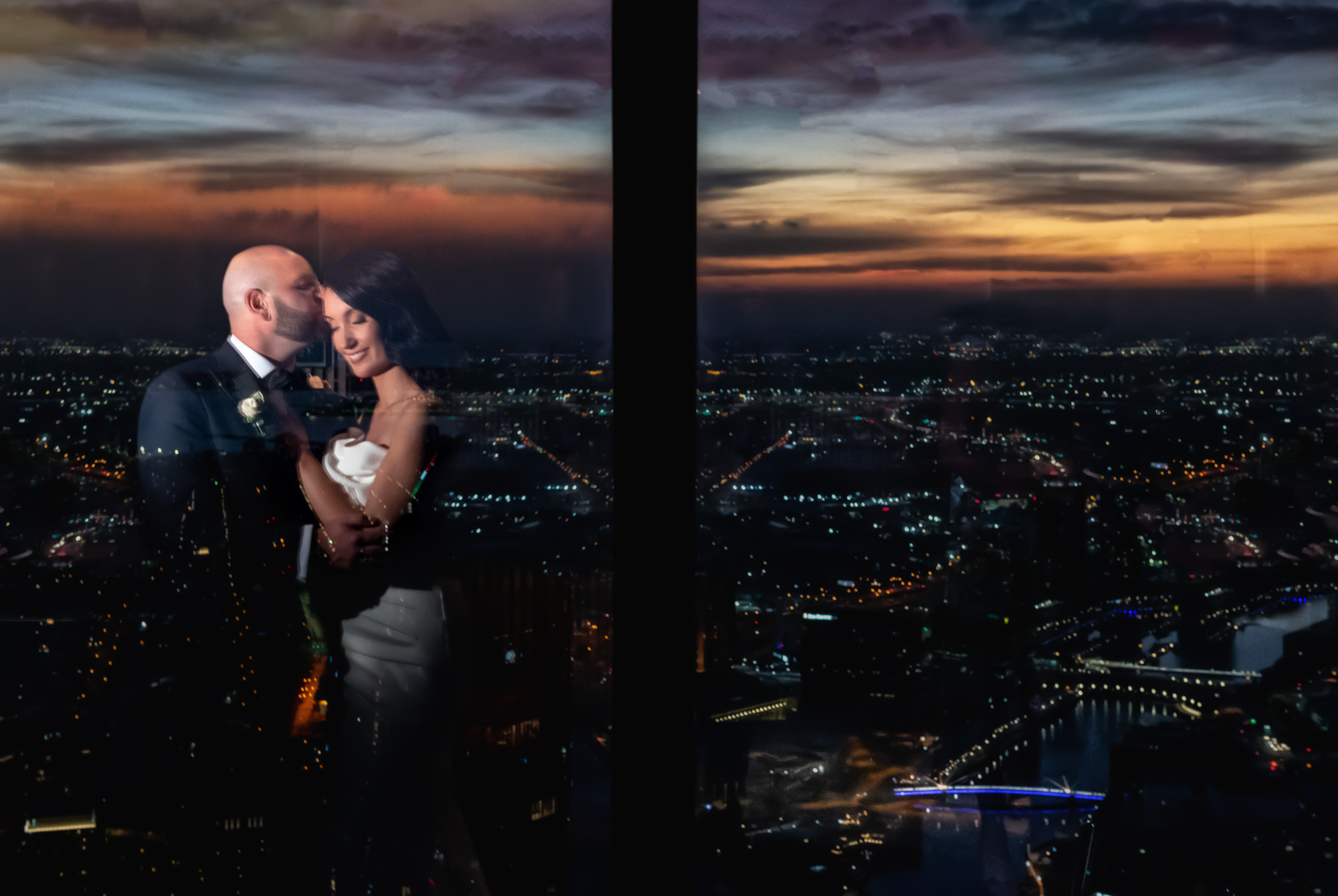 Bride and groom embrace by a window at night with a glowing city skyline and sunset reflected around them.