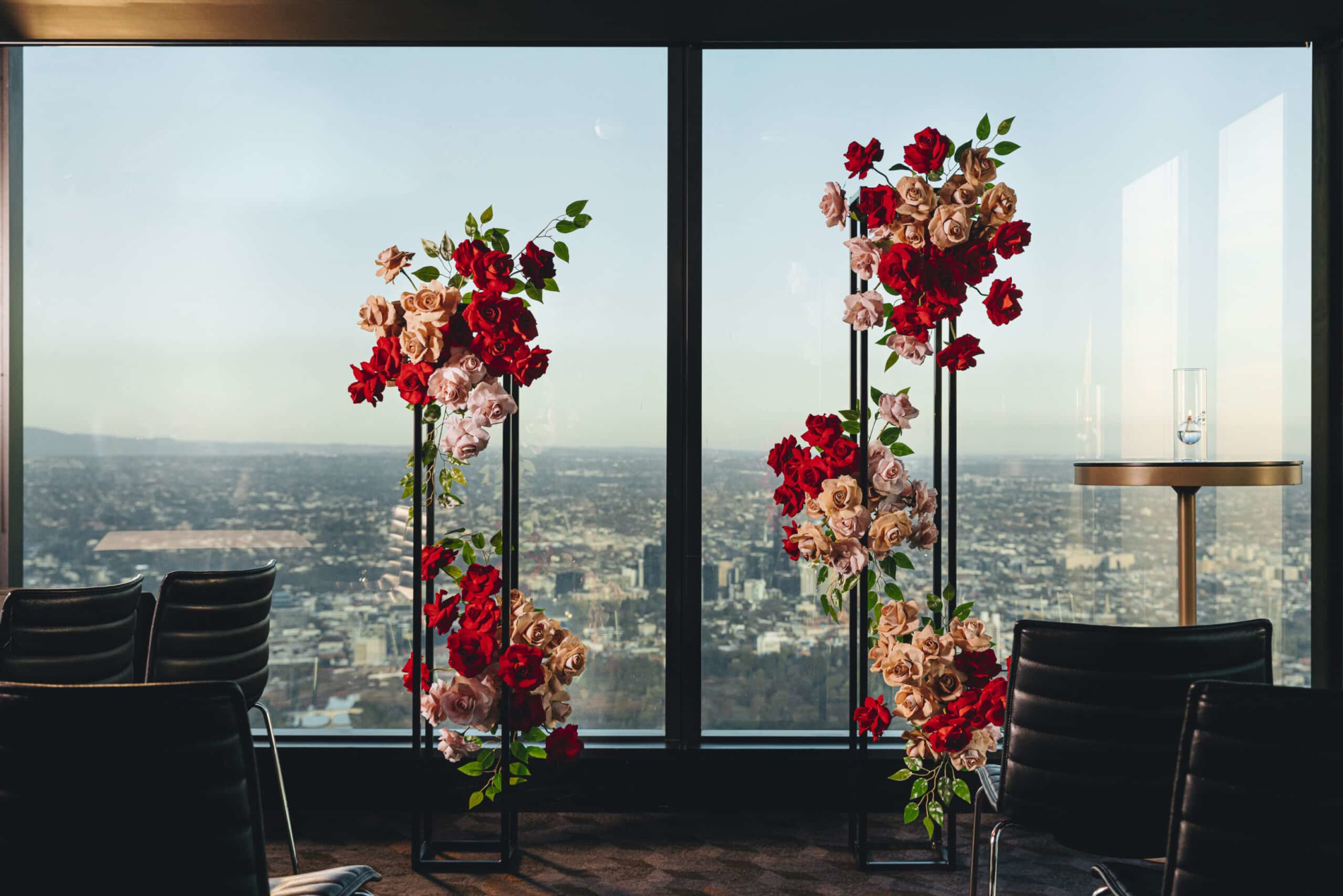 Modern indoor wedding ceremony setup with red and blush rose pillars against a panoramic city skyline view.