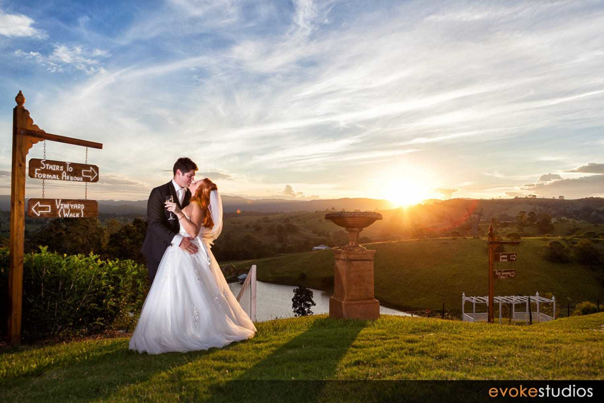Bride and groom embrace on a grassy hill at sunset overlooking a countryside lake and wedding arbour.
