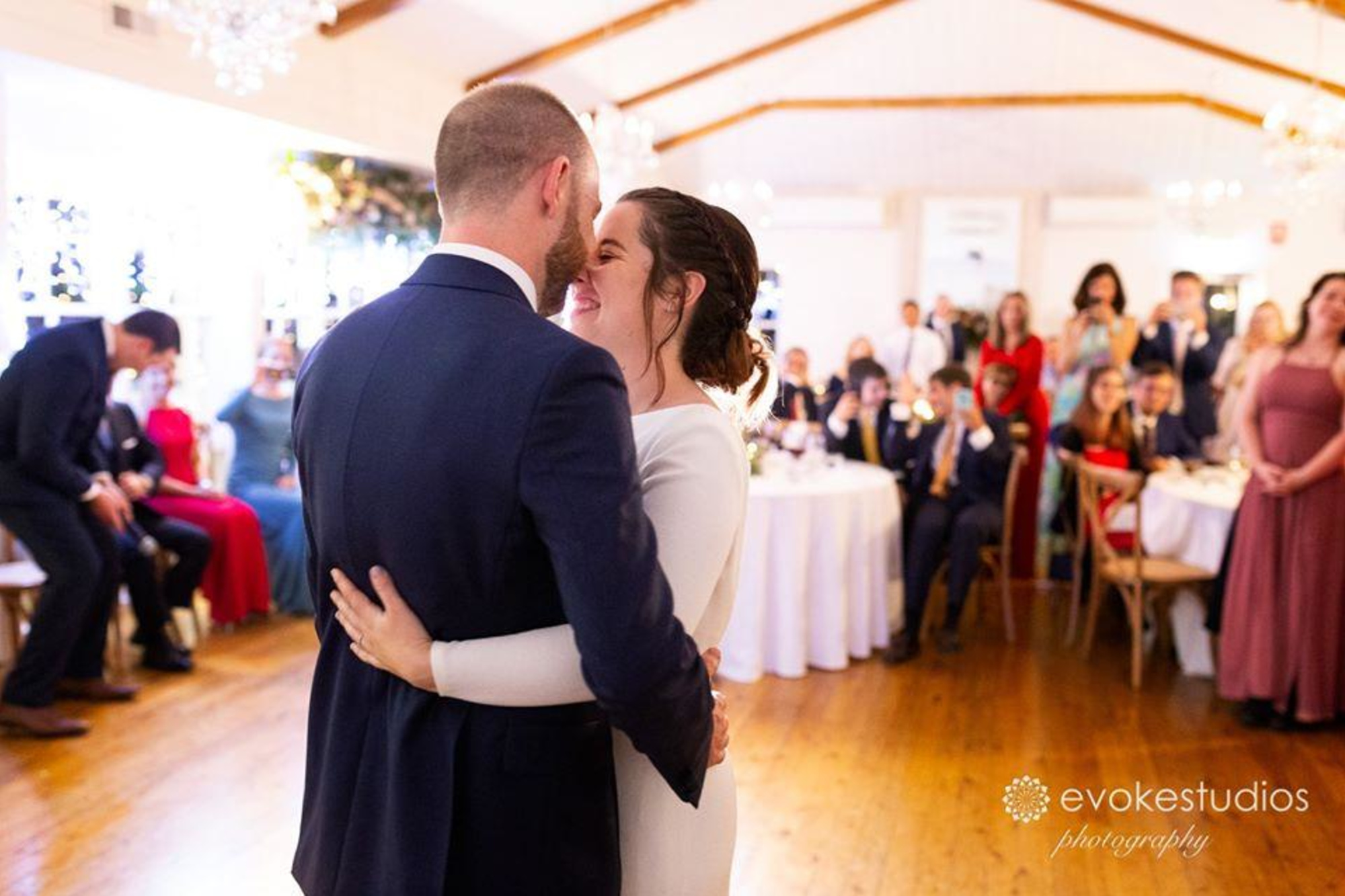 Bride and groom share a joyful first dance in a bright indoor reception surrounded by cheering guests.
