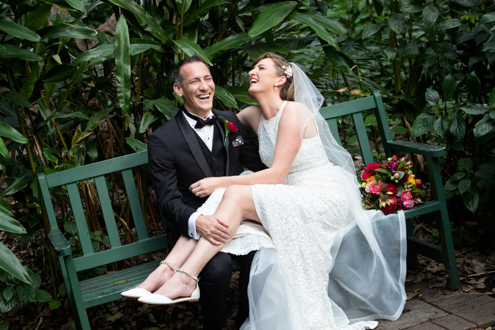 Laughing bride and groom sit on a green bench in a lush garden with a colorful bouquet beside them.