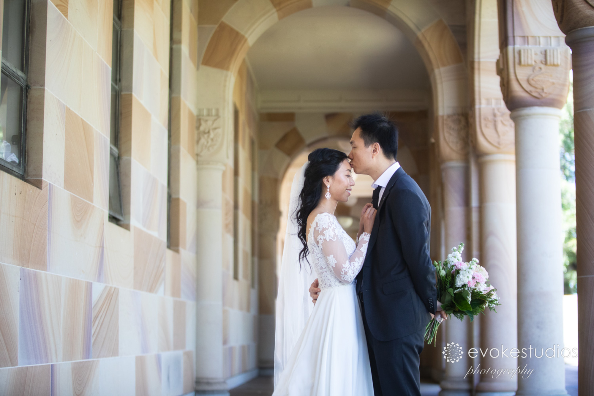 A groom kisses his bride's forehead as they embrace under stone arches, holding a pastel wedding bouquet.