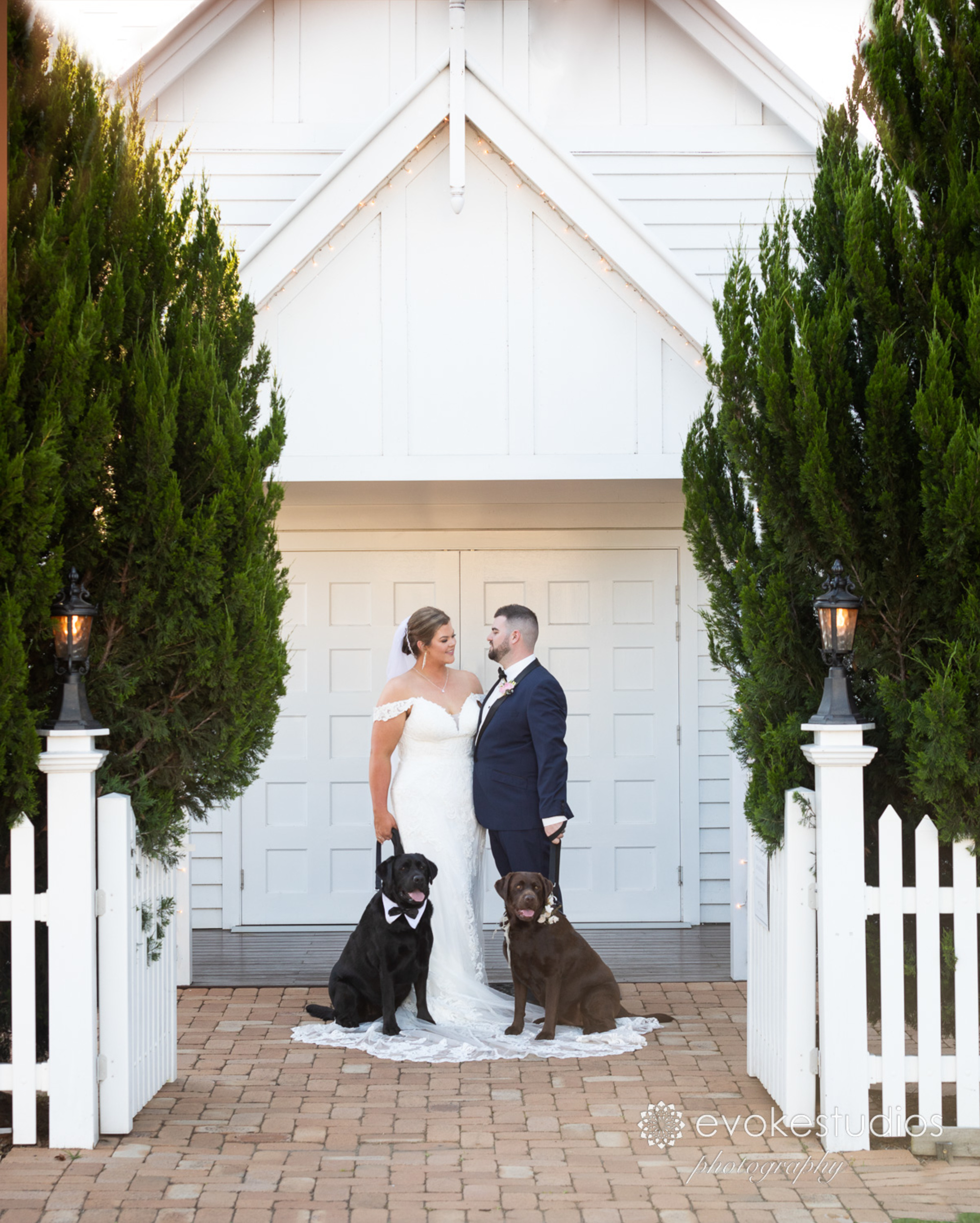 Bride and groom stand with two dogs in front of a white chapel framed by tall greenery.