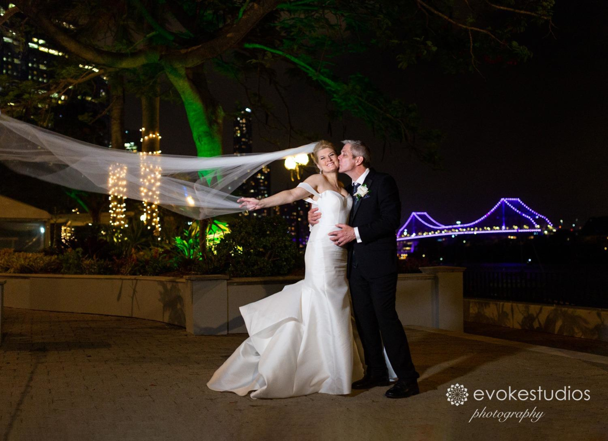 Bride and groom embrace at night with flowing veil and illuminated city bridge in the background.