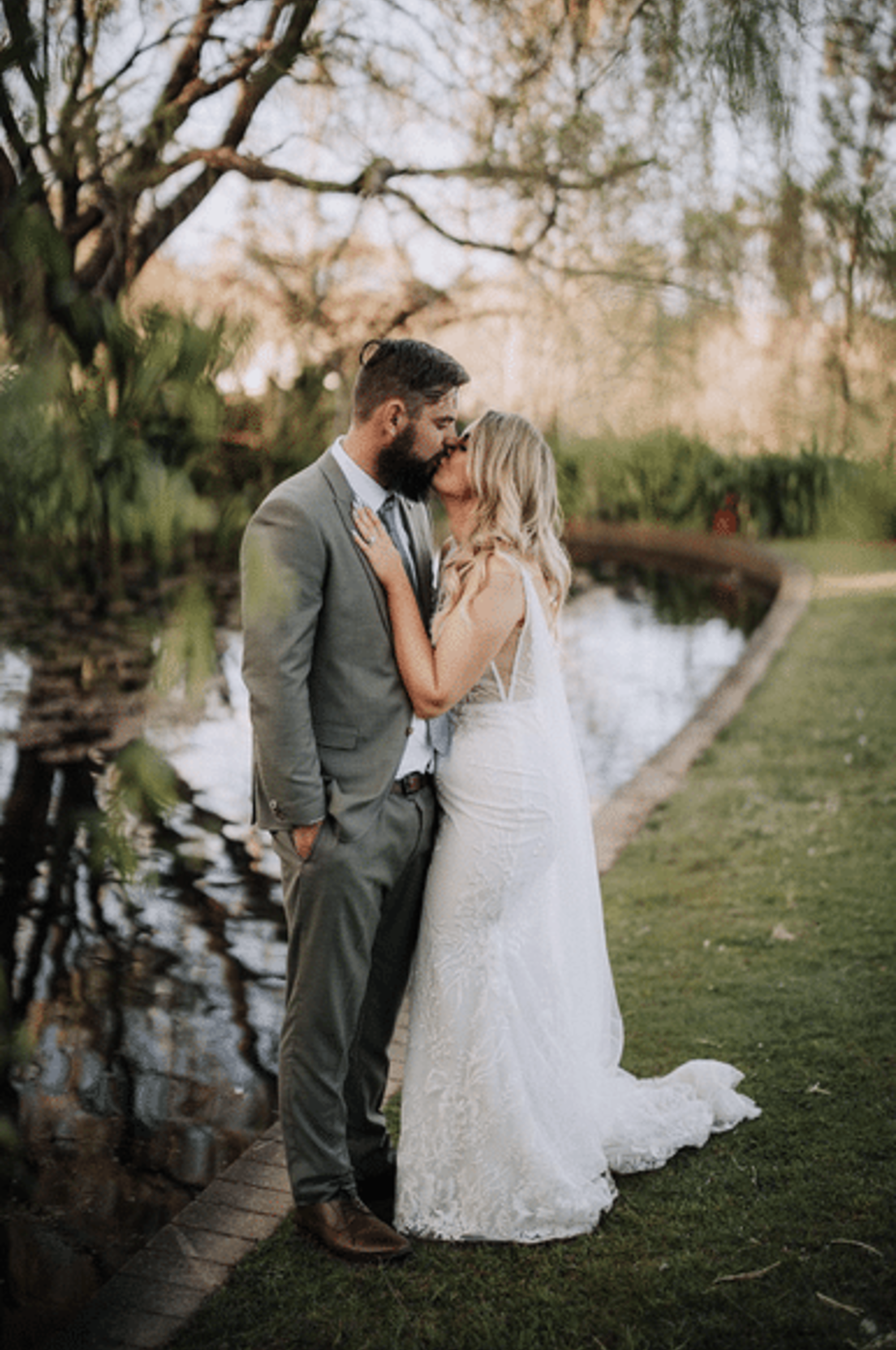 Bride and groom share an intimate kiss beside a serene pond in a lush garden setting.