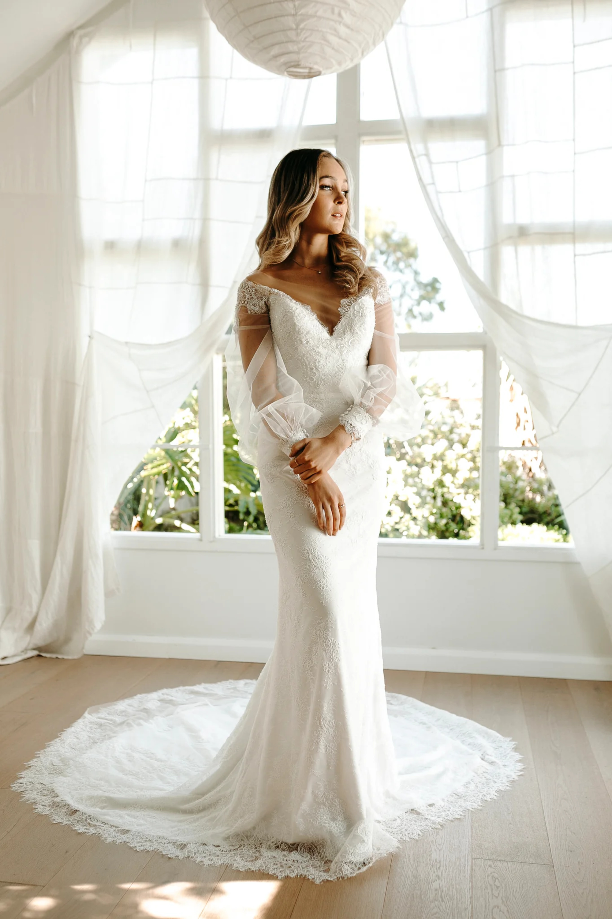 Bride in a fitted lace wedding gown with sheer sleeves standing in a bright room with flowing white curtains.
