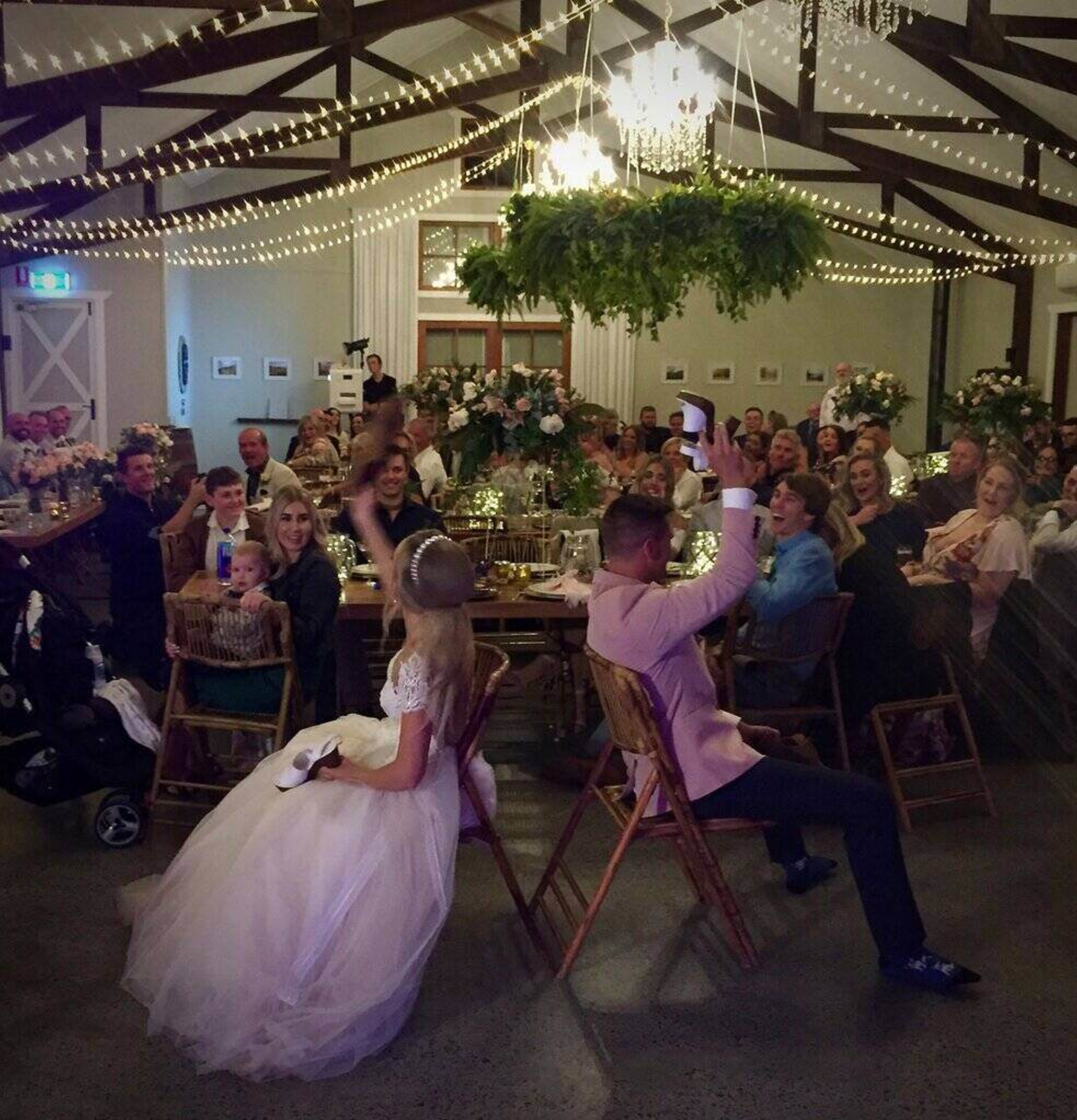 Indoor wedding reception with guests watching the couple play a shoe game under string lights and greenery decor.