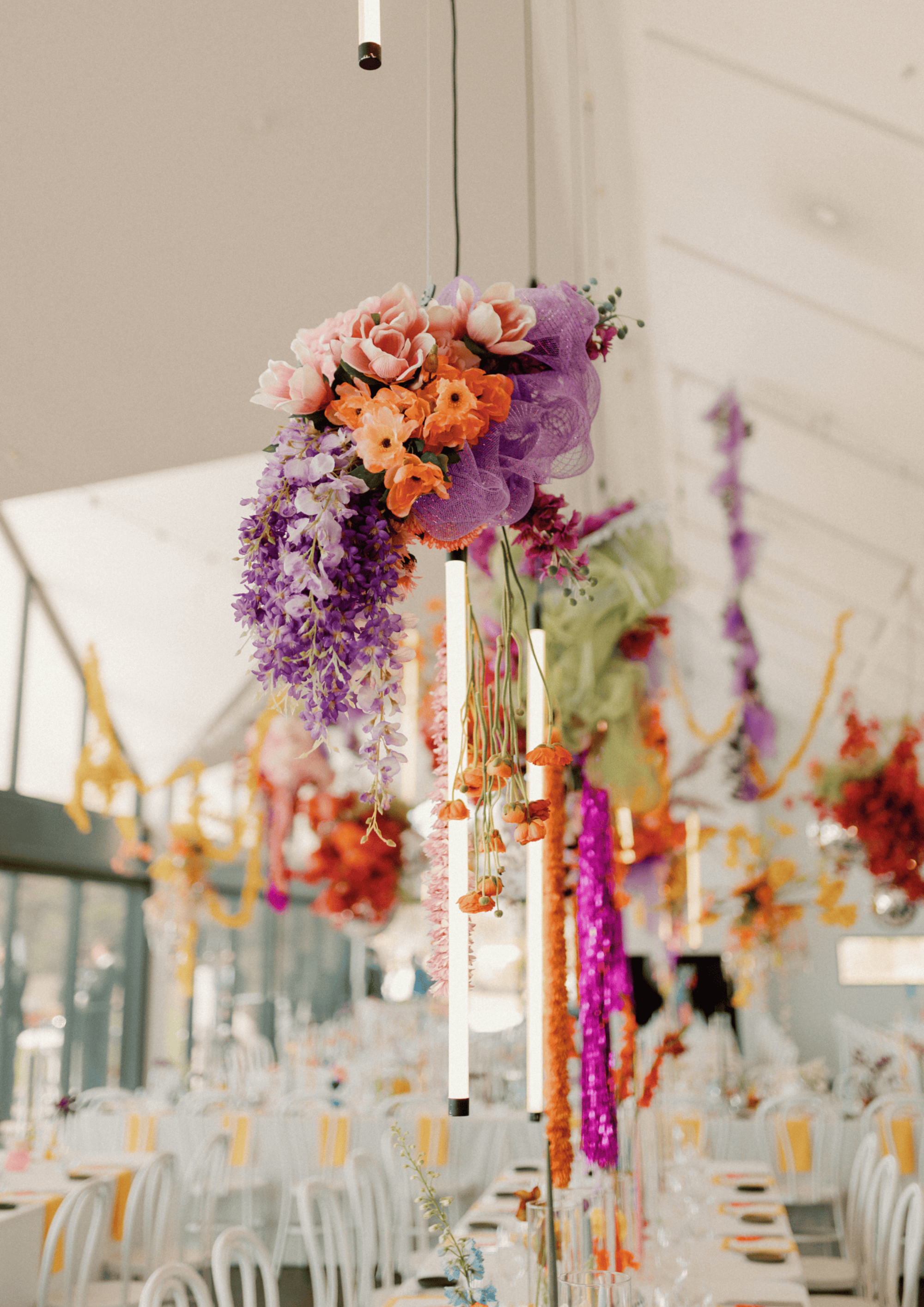 Colorful hanging floral installations decorate a bright indoor wedding reception with long banquet tables below.