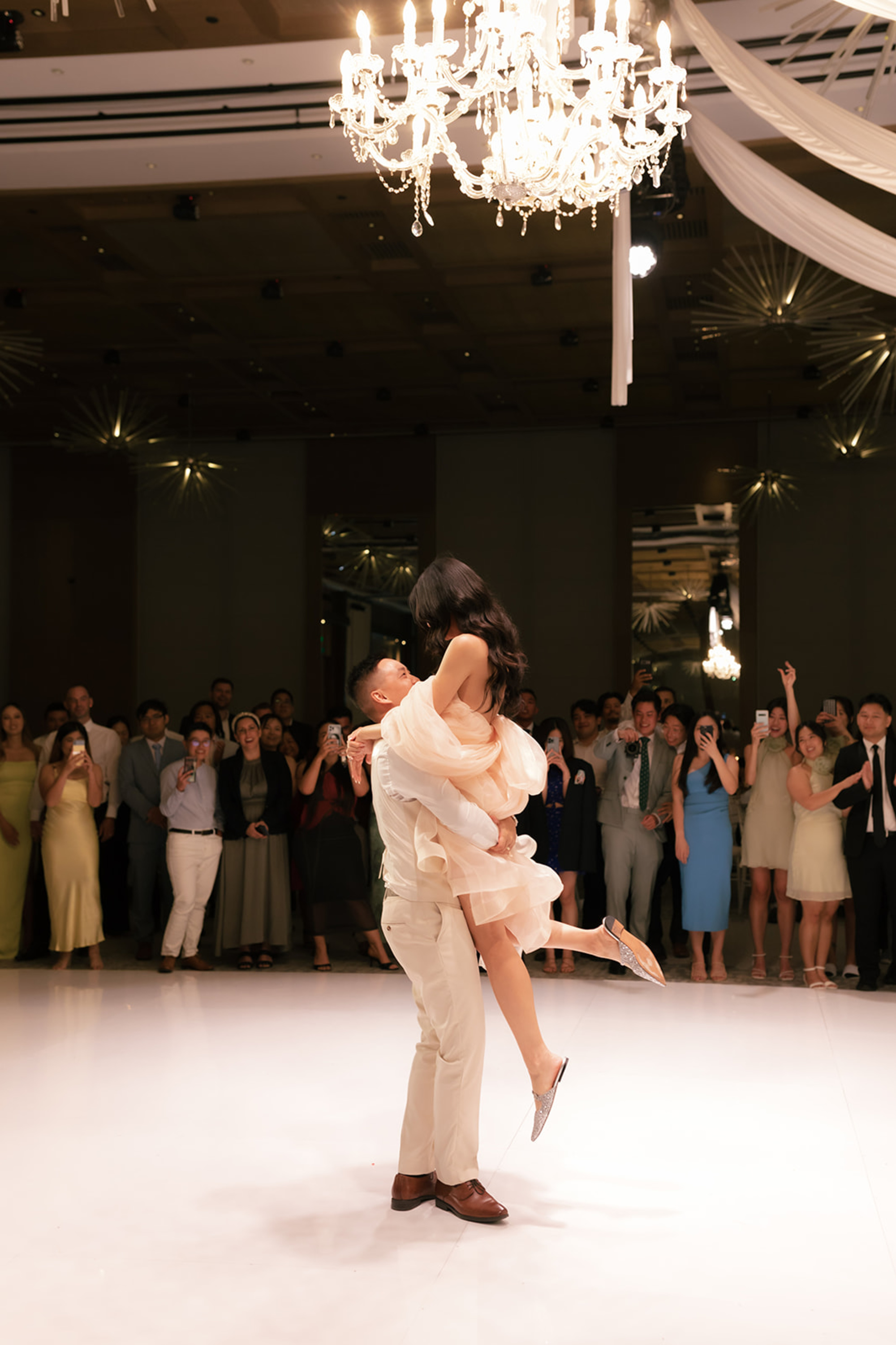 A groom lifts his bride on the dance floor under a chandelier while guests watch and take photos.