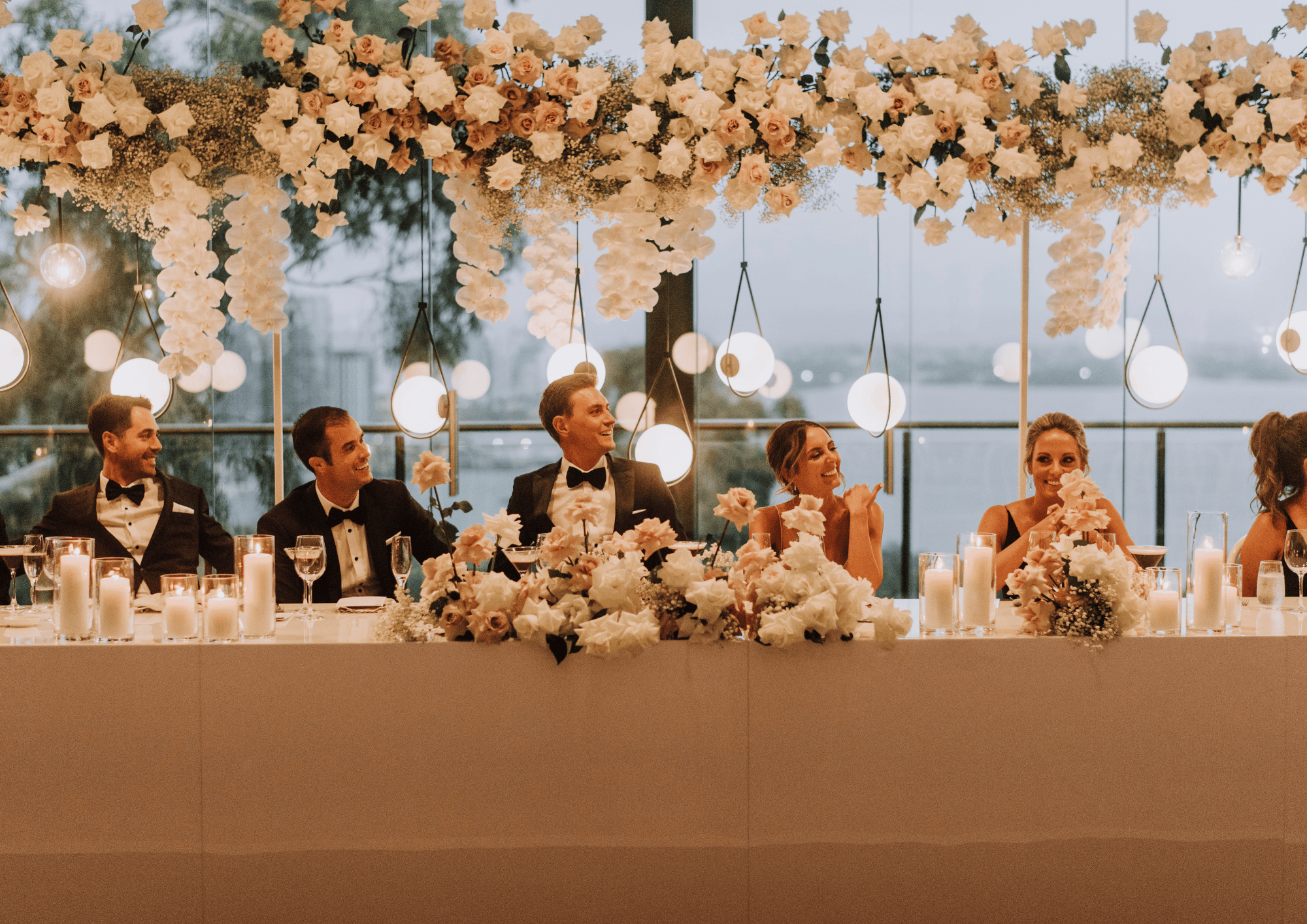Elegant wedding head table with bridal party seated beneath lush floral installations and warm hanging lights.