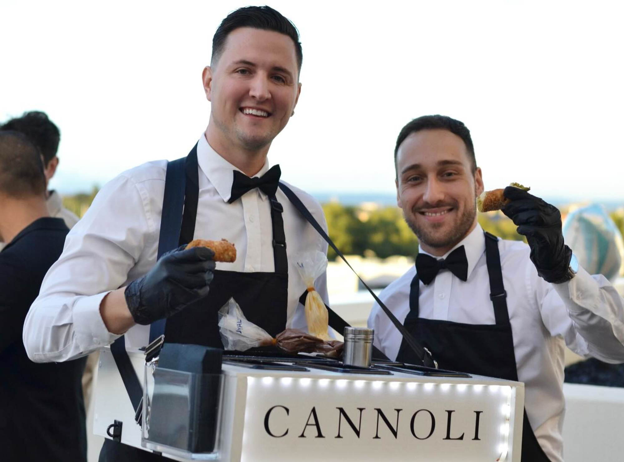 Two smiling servers in bow ties offer cannoli from a mobile dessert cart at an outdoor wedding reception.