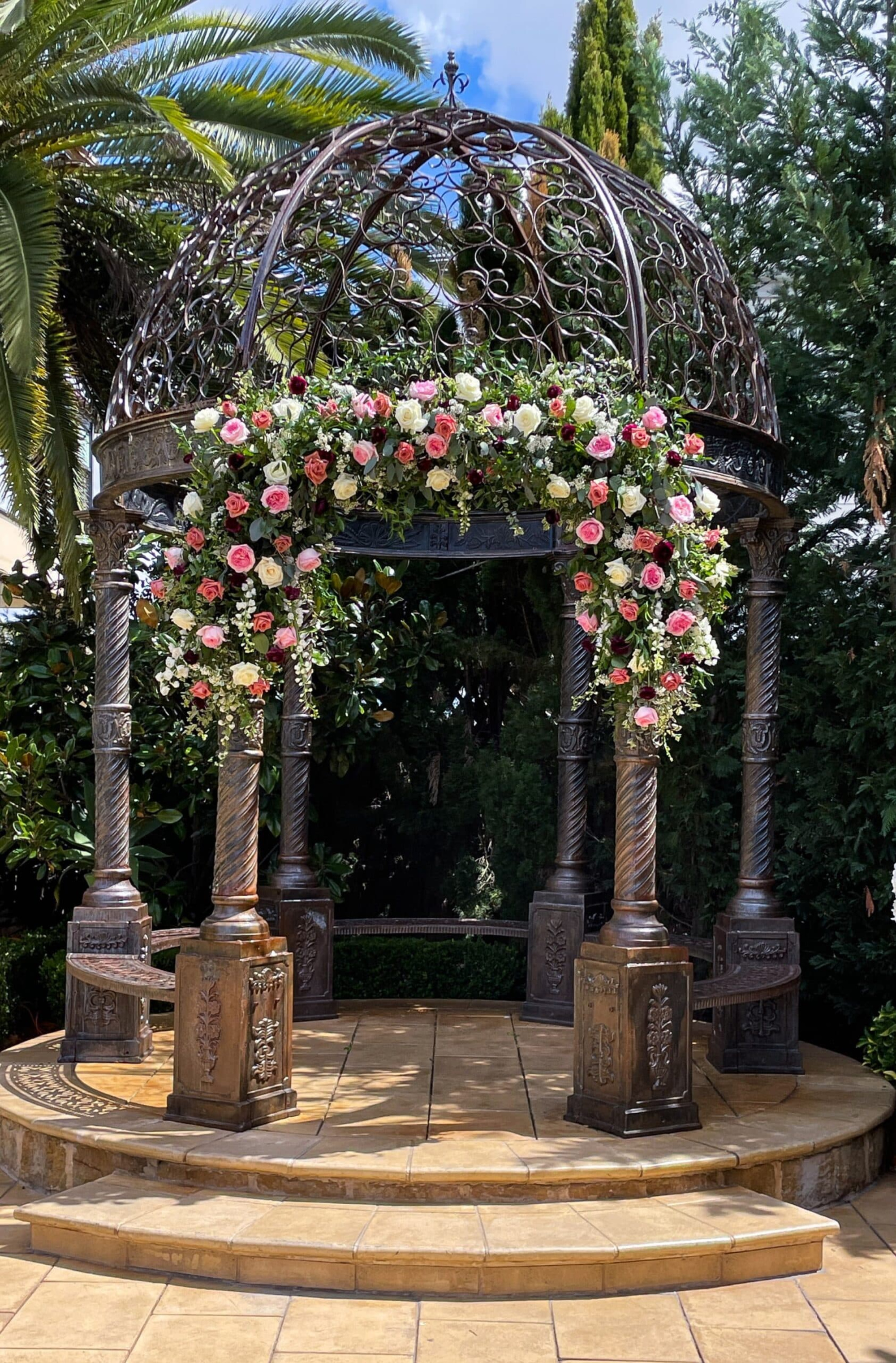 Ornate outdoor gazebo decorated with cascading pink and white roses for a romantic garden wedding ceremony.