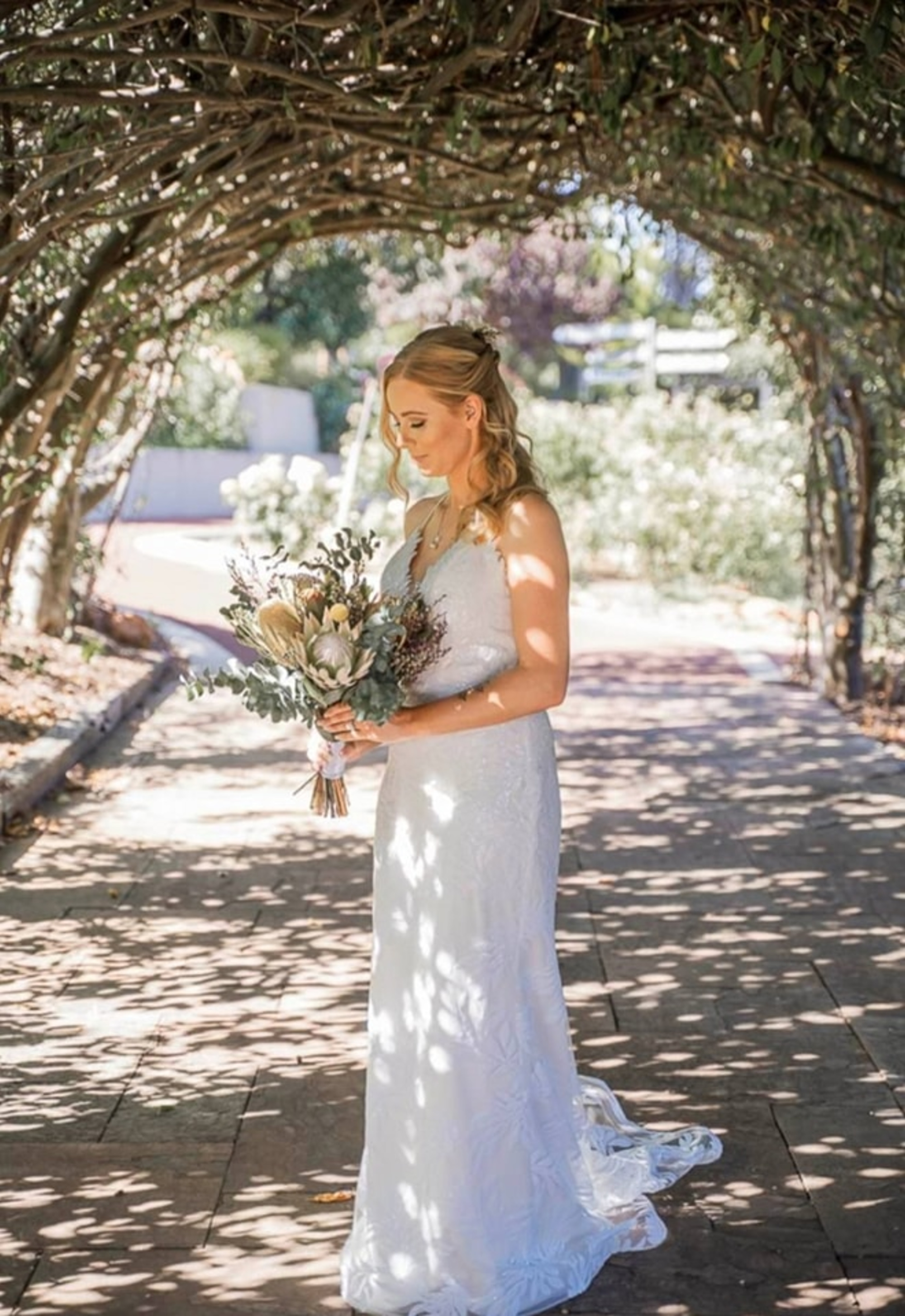Bride in a lace gown holding a rustic bouquet beneath a leafy garden archway with dappled sunlight.