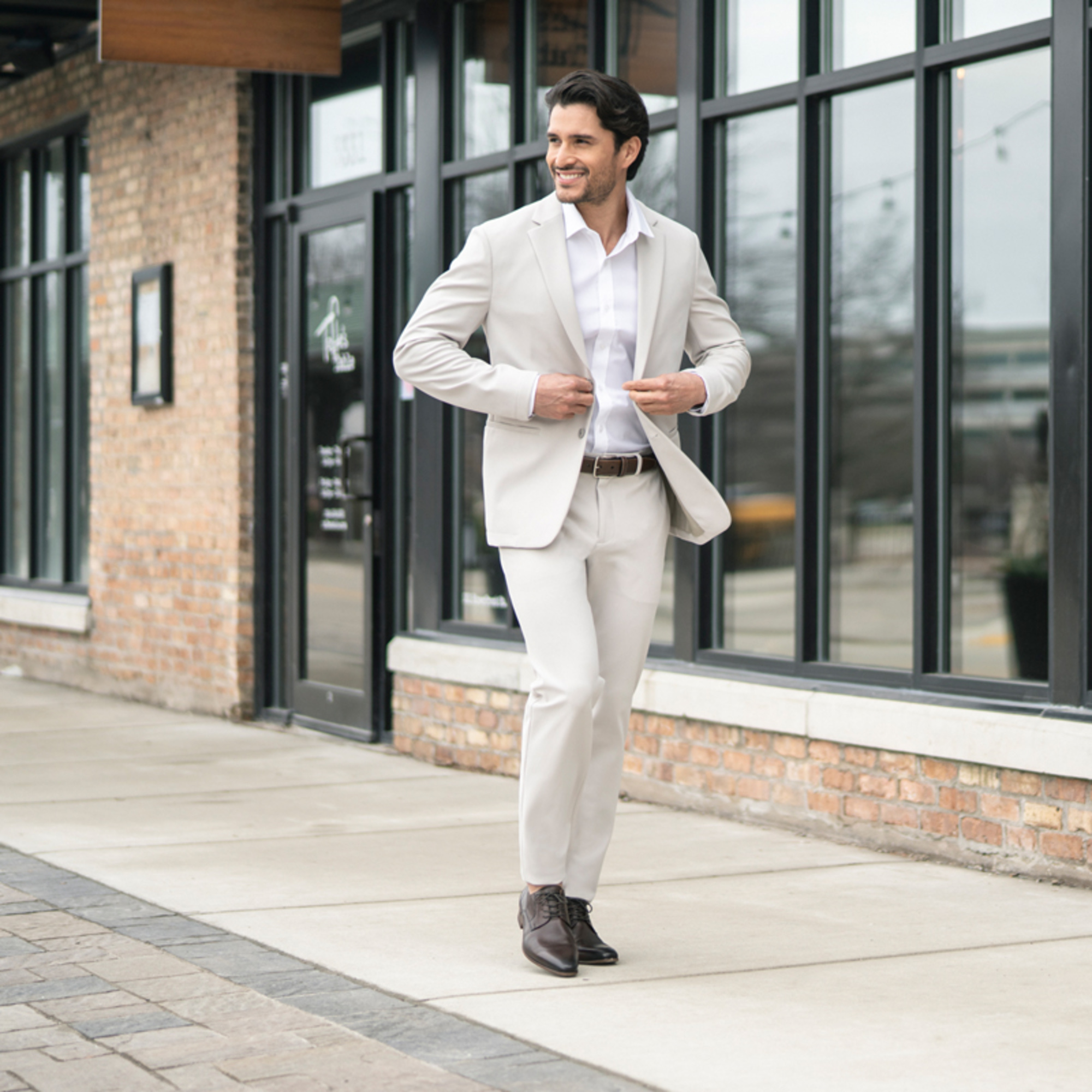 Smiling man in a light beige suit walking on a city sidewalk in front of large windows.