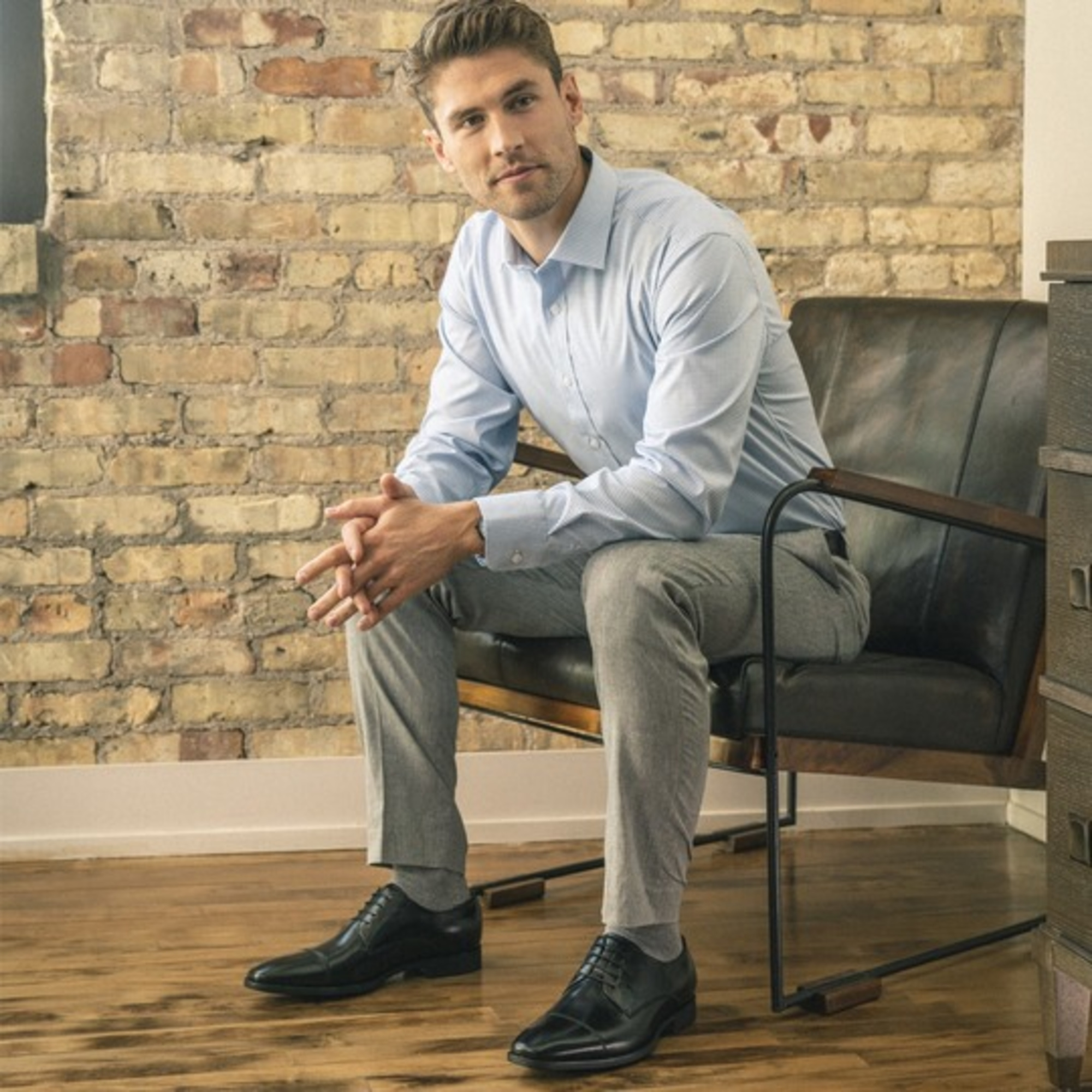 Well-dressed man in a light blue shirt and gray trousers sits on a leather chair against an exposed brick wall.