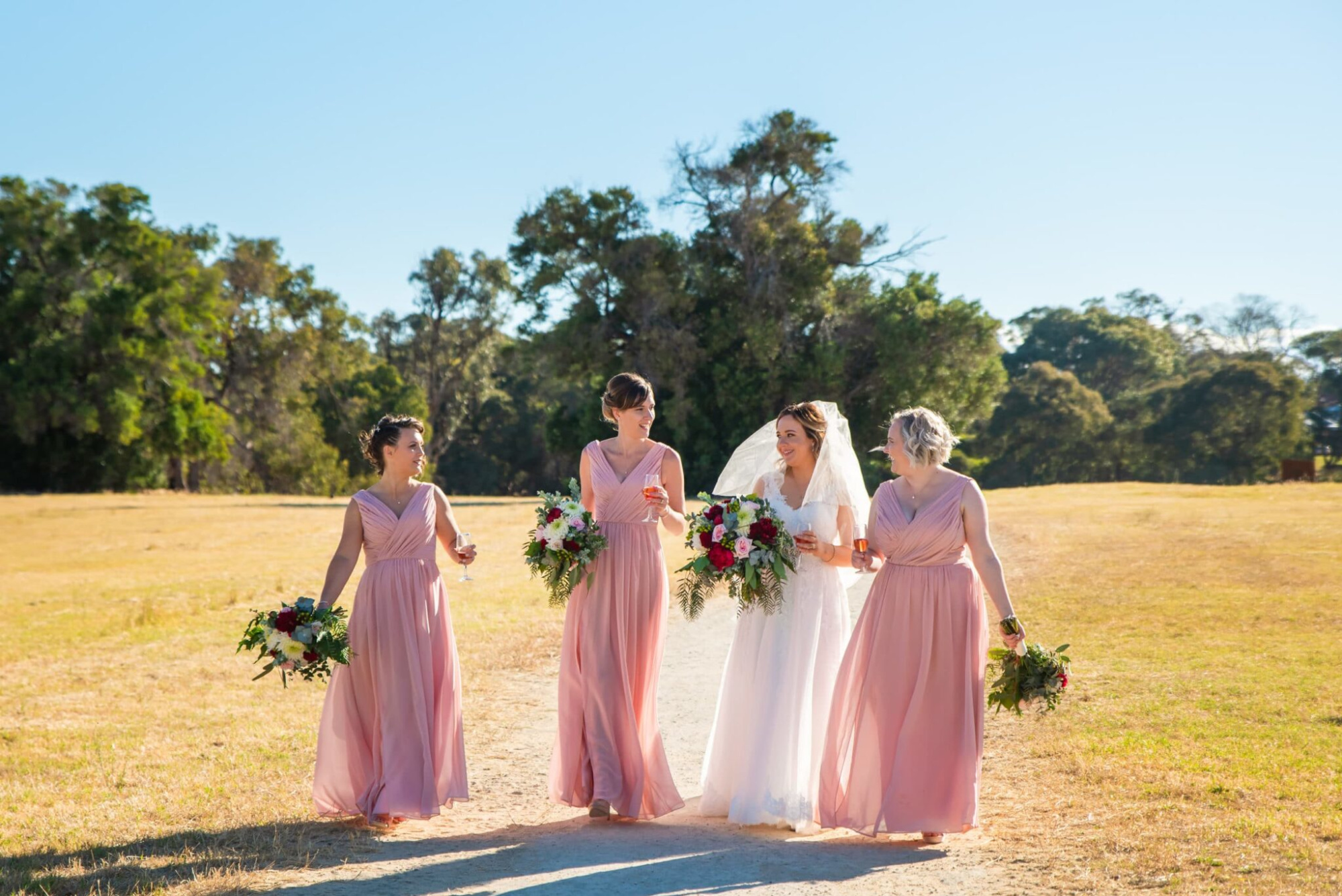 Bride and bridesmaids in pink dresses walking outdoors with bouquets on a sunny wedding day.