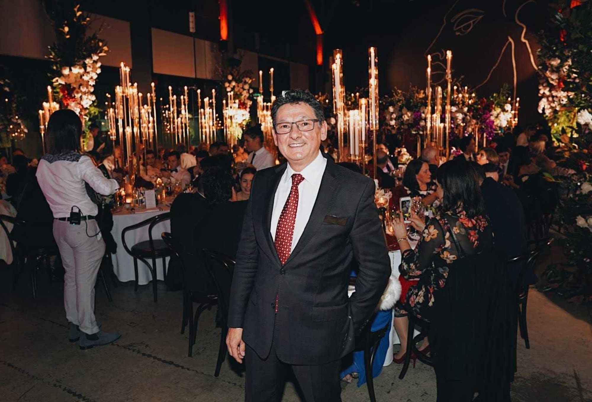 Man in a suit stands smiling at an elegant candlelit wedding reception with seated guests behind him.