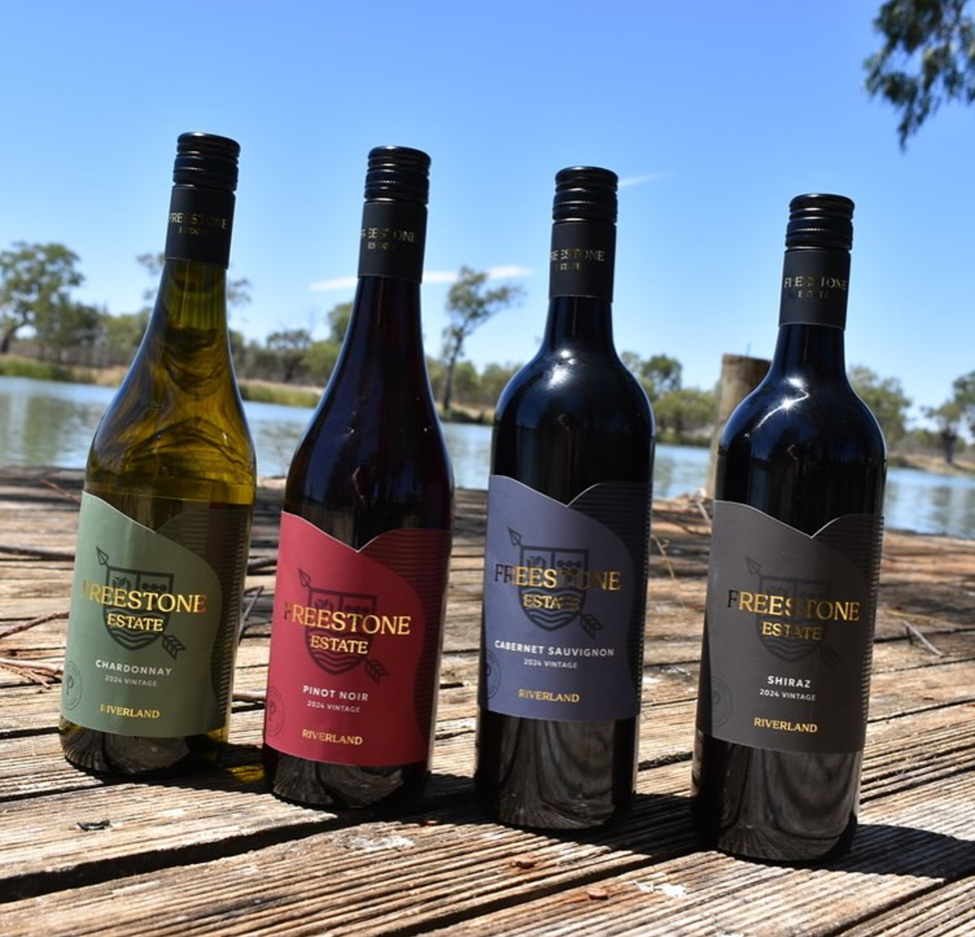 Four Freestone Estate wine bottles displayed on wooden decking beside a riverside backdrop.