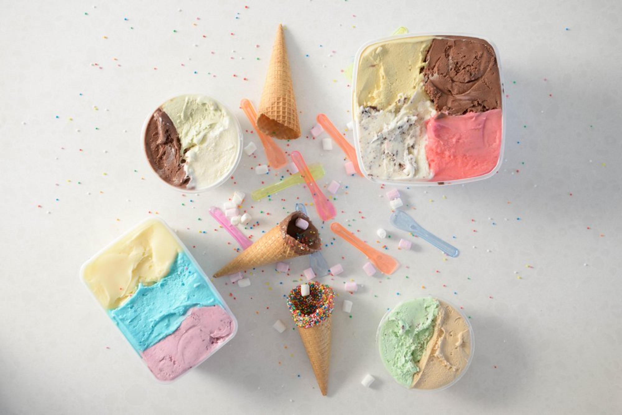 Colorful tubs of ice cream with waffle cones, sprinkles, and spoons arranged on a light surface.