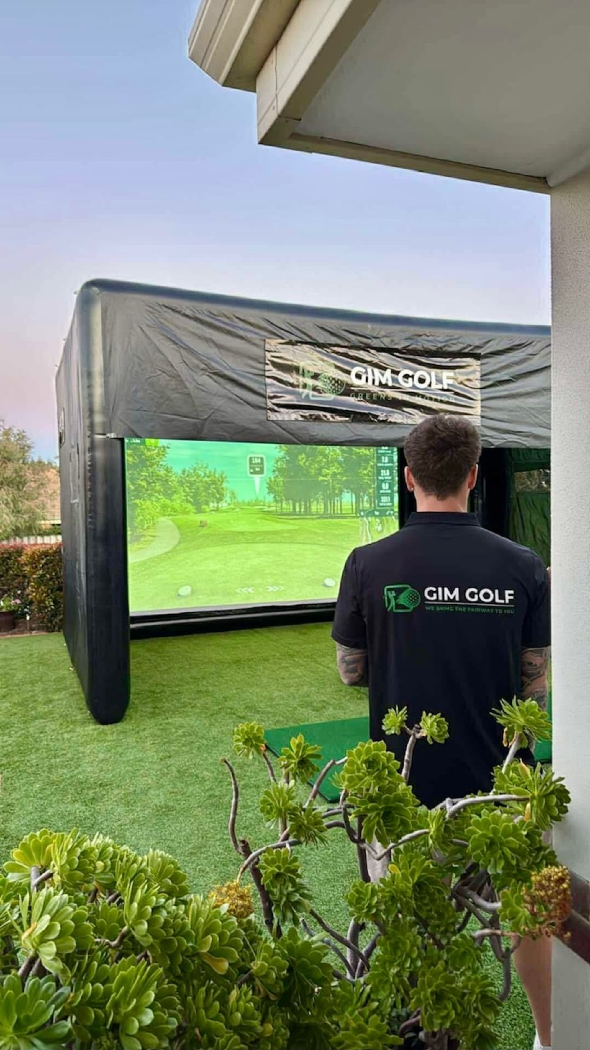 Man using an outdoor golf simulator setup on artificial grass, branded with GIM Golf signage.