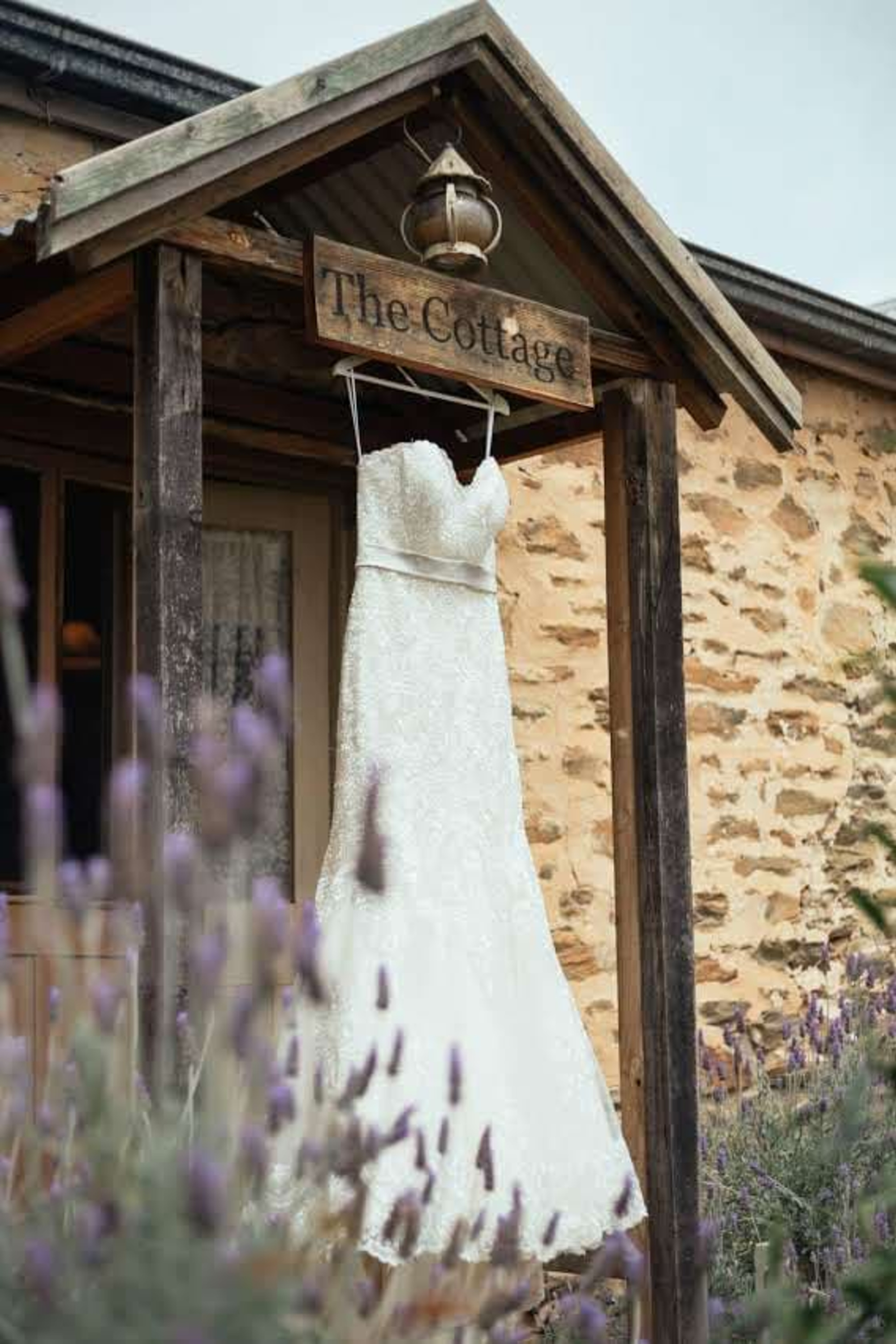 Lace wedding dress hanging outside a rustic stone cottage surrounded by lavender.