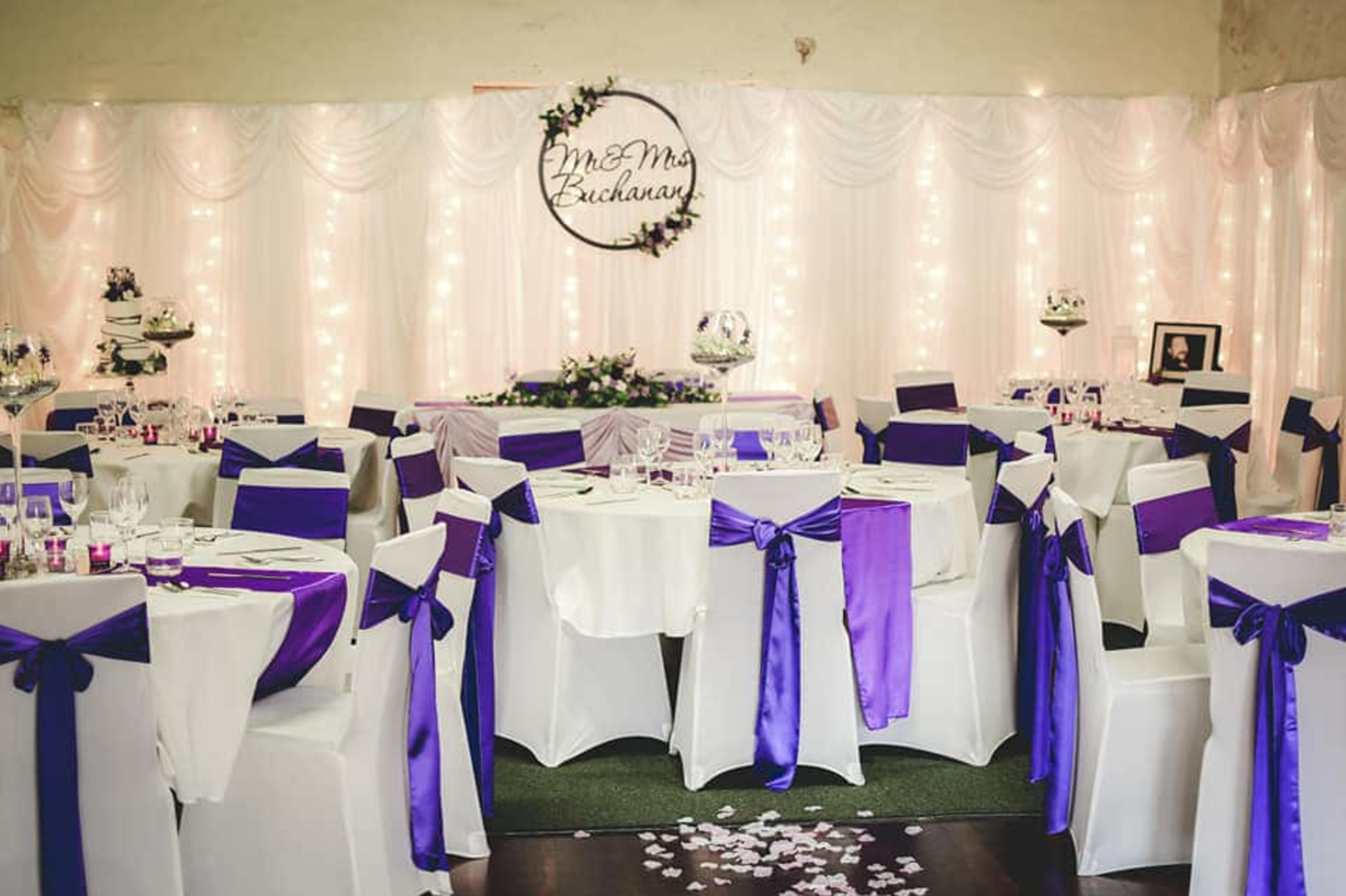 Elegant indoor wedding reception with purple chair sashes, round tables, and a draped backdrop with fairy lights.