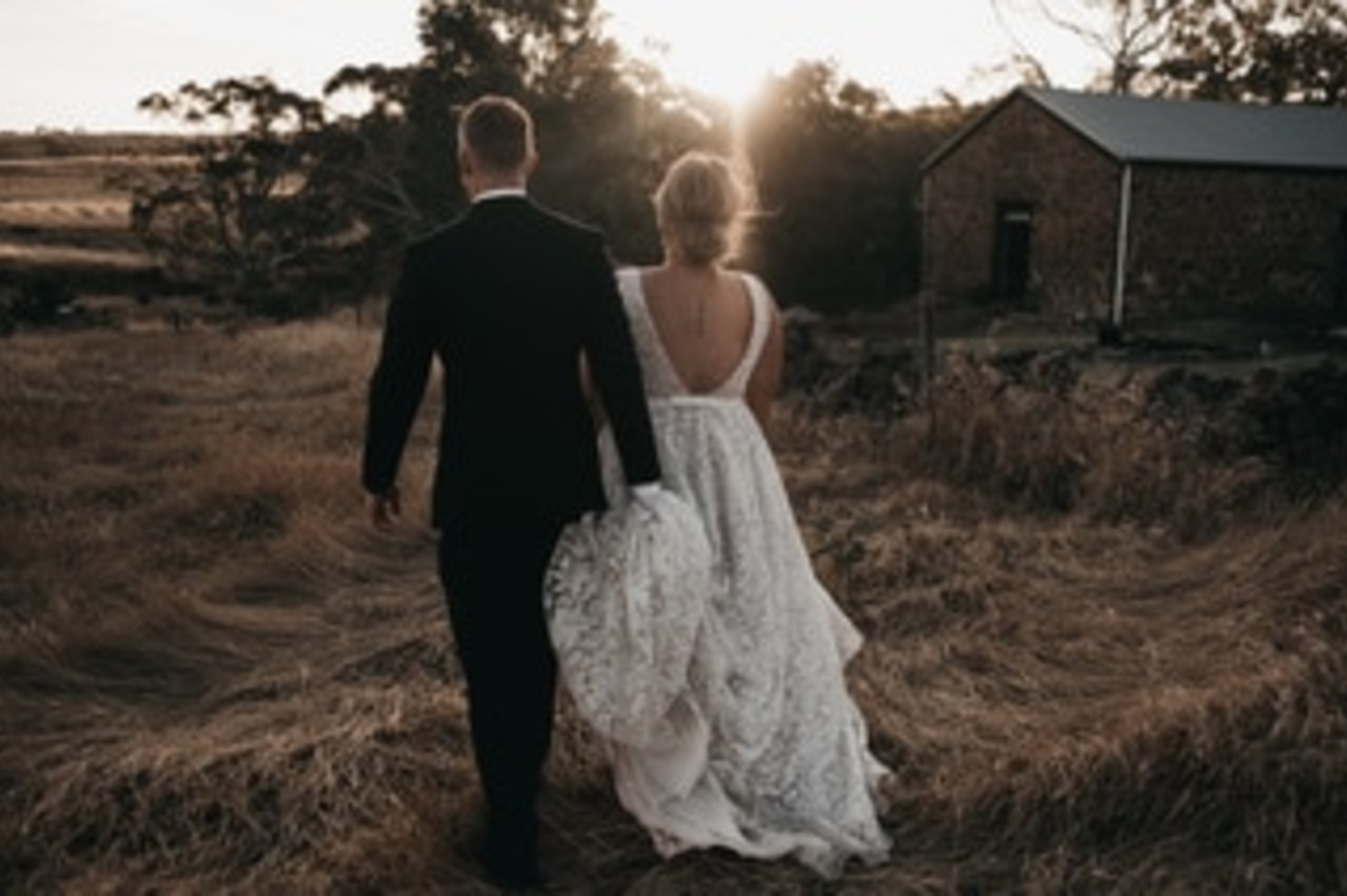 Bride and groom walk hand in hand through a rustic field at sunset near a stone barn.