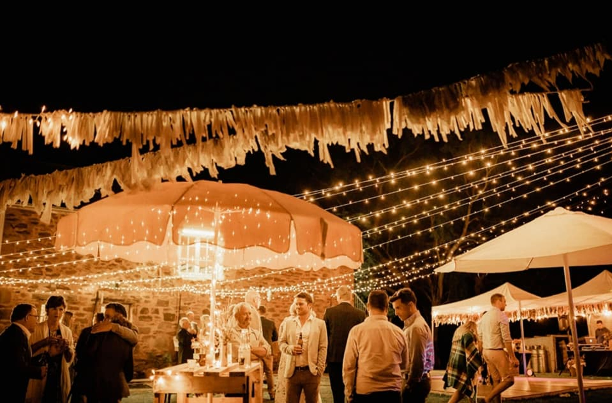 Outdoor evening wedding reception with guests mingling under string lights and festive umbrellas.