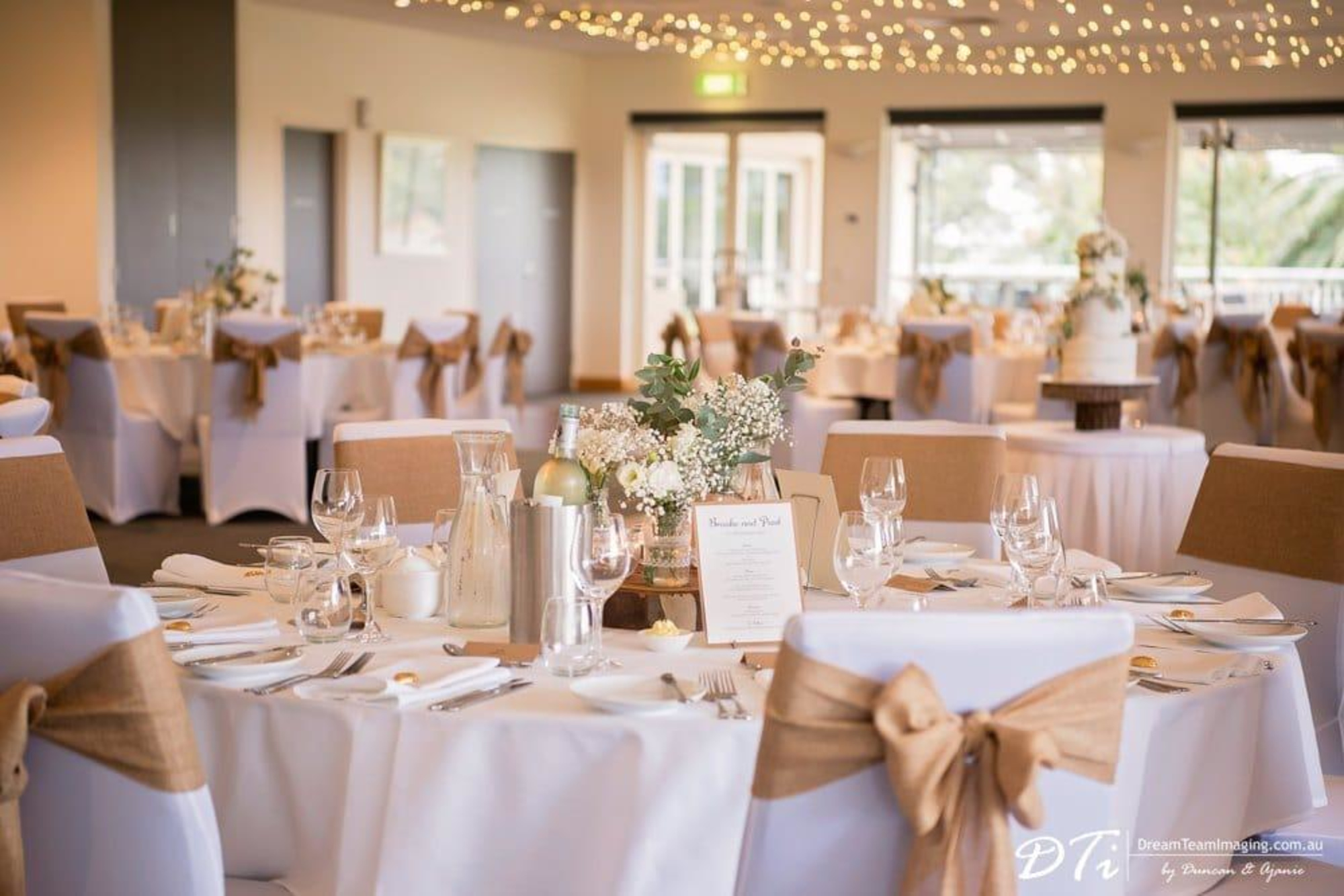 Rustic chic wedding reception room with round tables, burlap chair sashes, and soft string lights overhead.
