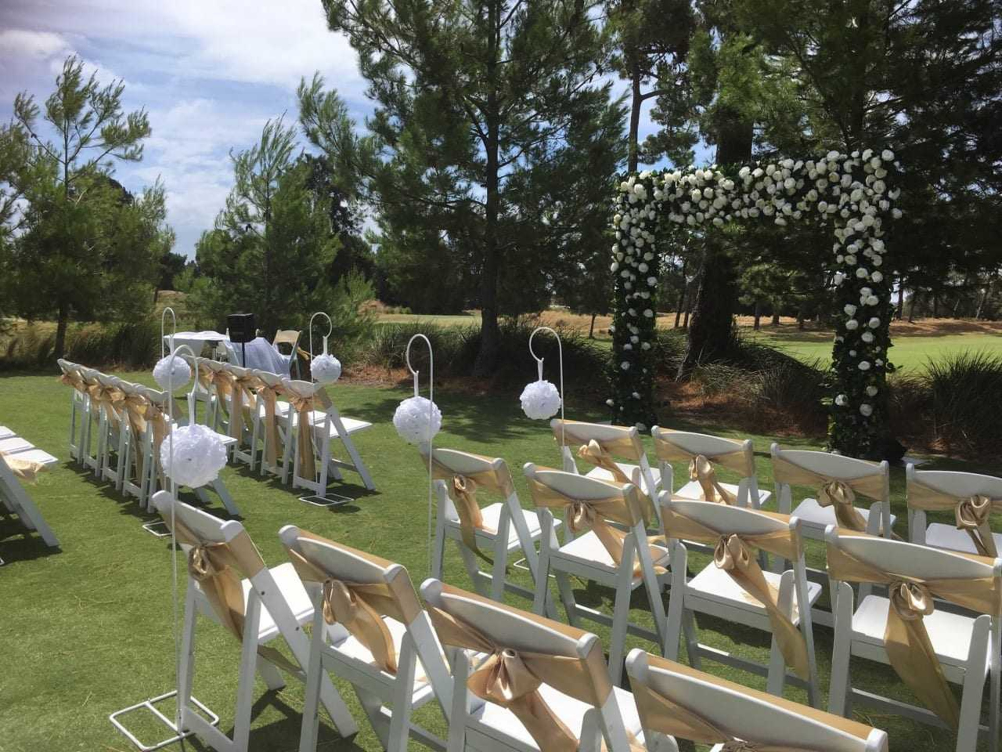 Outdoor wedding ceremony setup with white chairs, gold sashes, and a flower-covered arch on a grassy lawn.