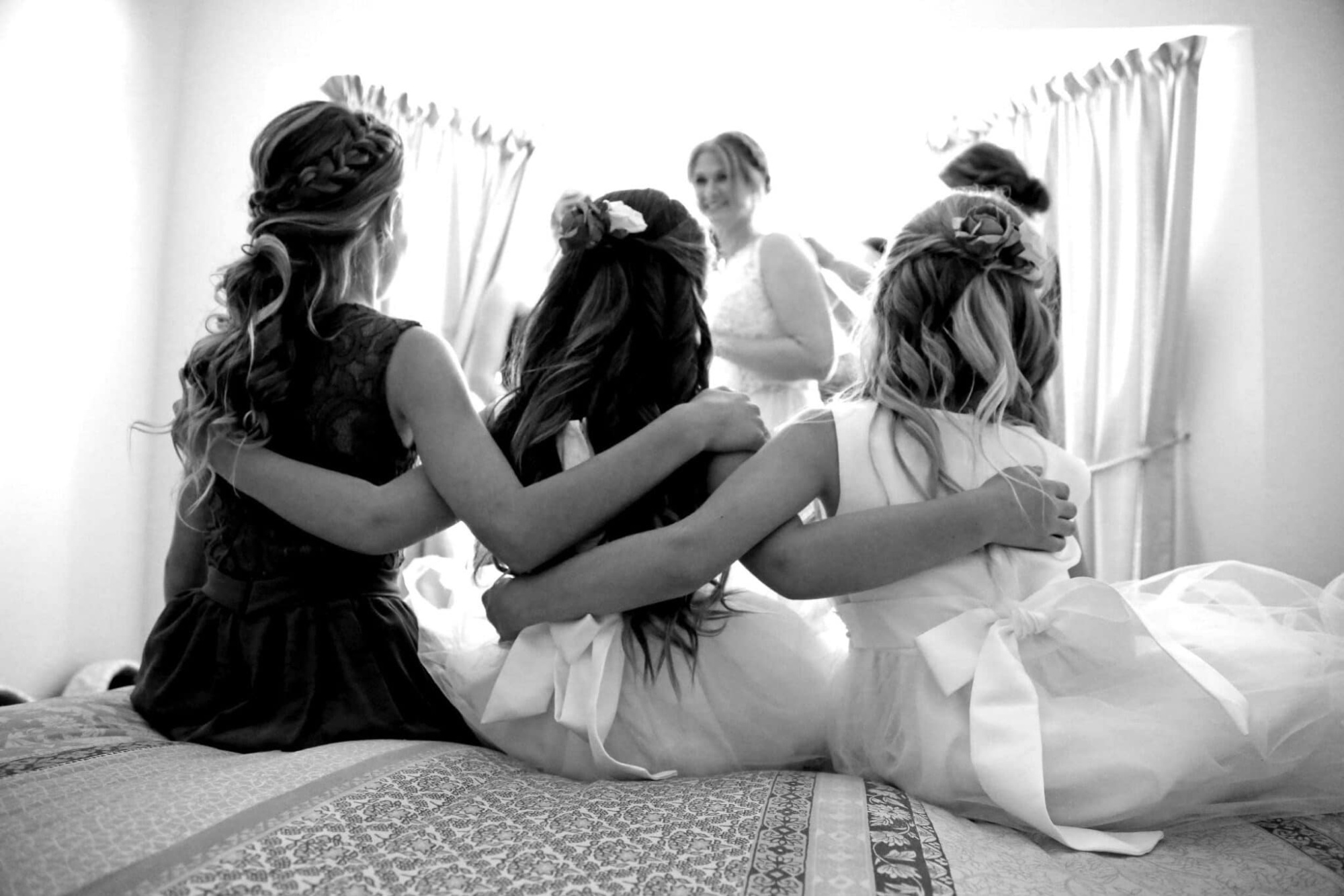 Three girls with linked arms sit on a bed watching the bride get ready in a bright room.