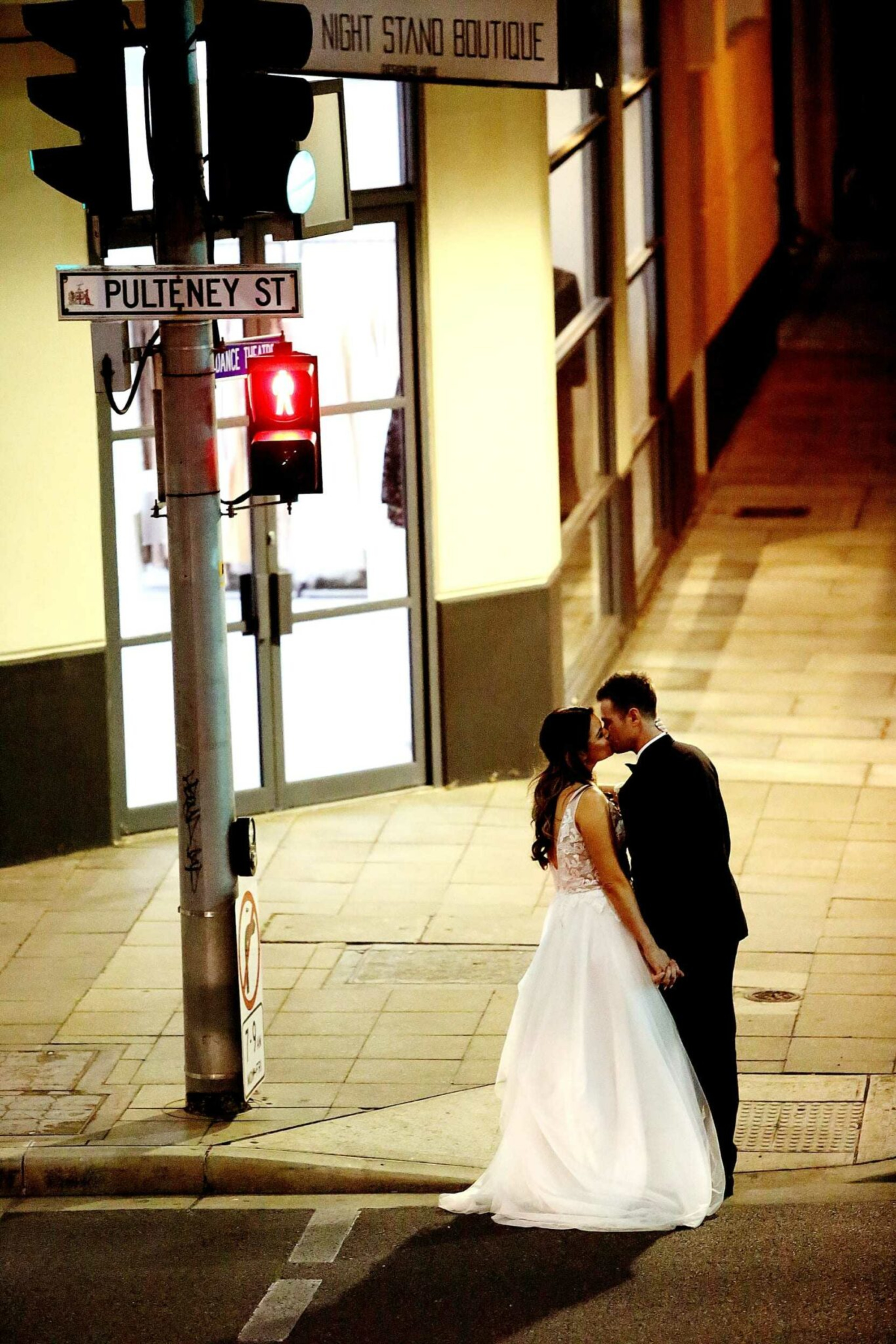 Bride and groom share a kiss at night on a city street corner beneath a traffic light.