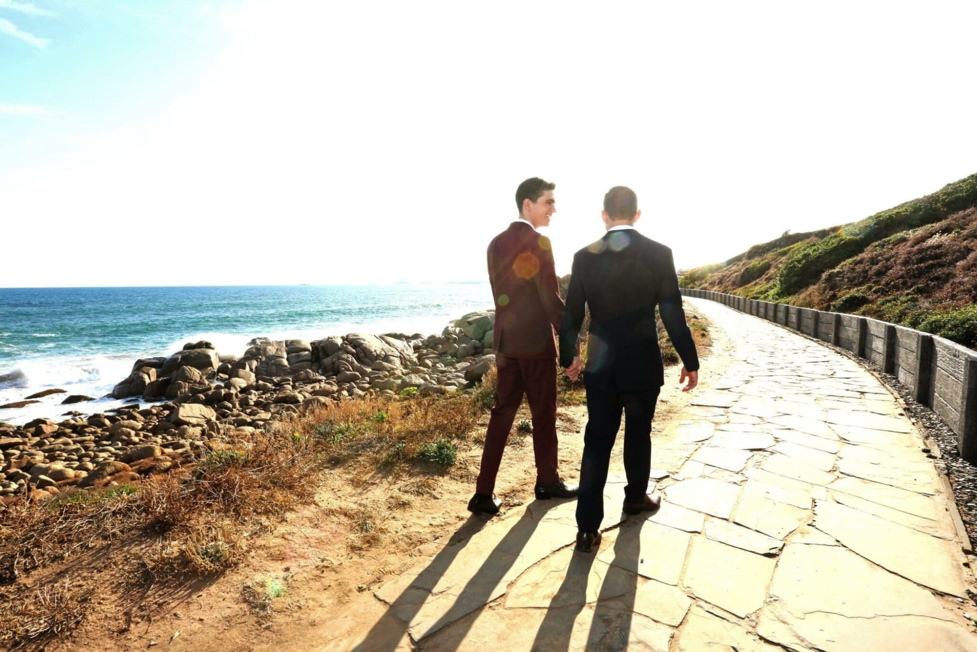 Two grooms in suits walk hand in hand along a sunny coastal path by the ocean.