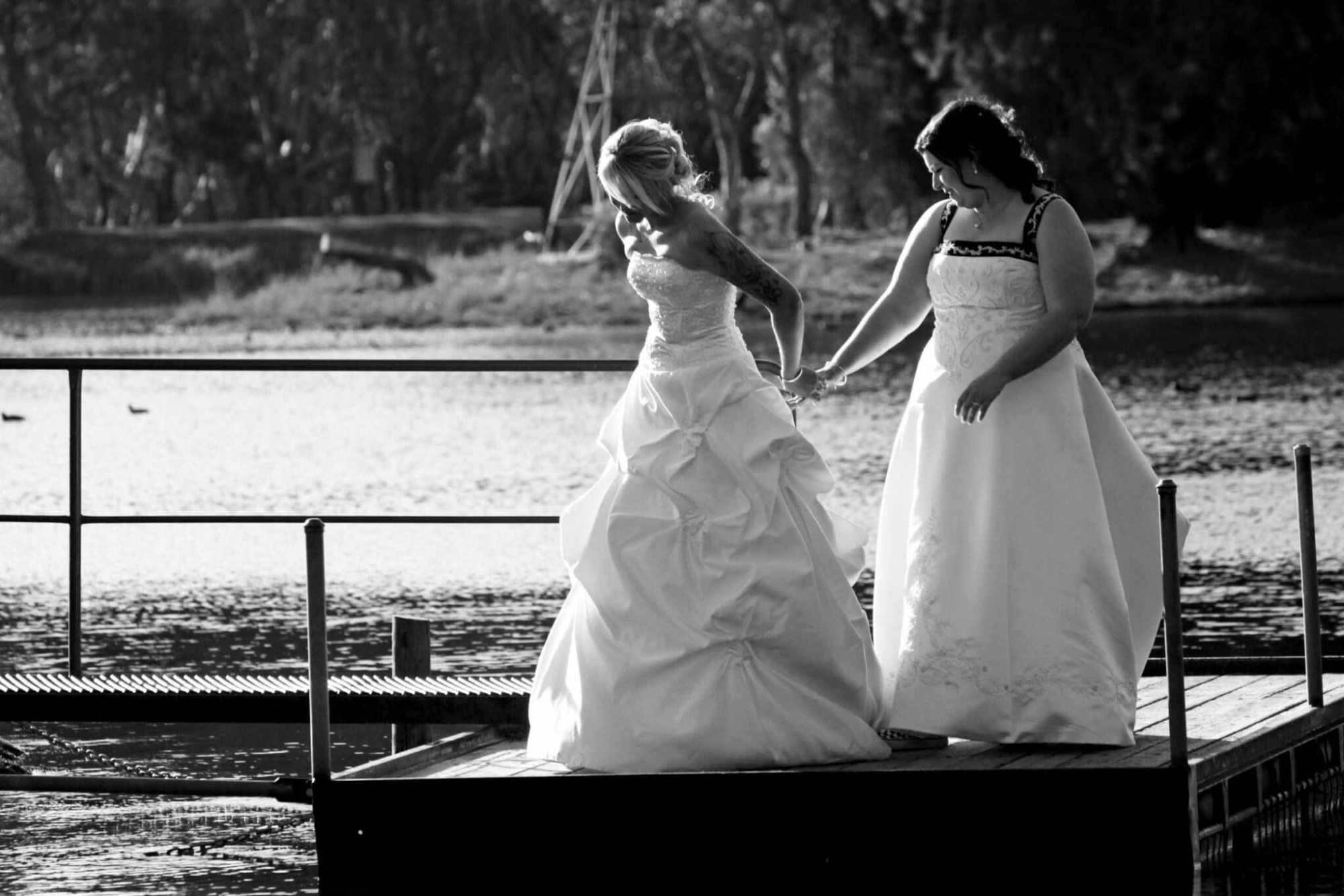 Two brides in wedding gowns hold hands on a lakeside dock in soft black and white light.