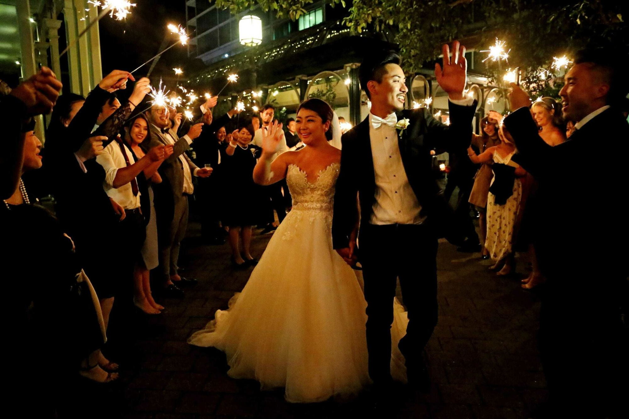 Bride and groom walk through a nighttime sparkler exit surrounded by cheering wedding guests.
