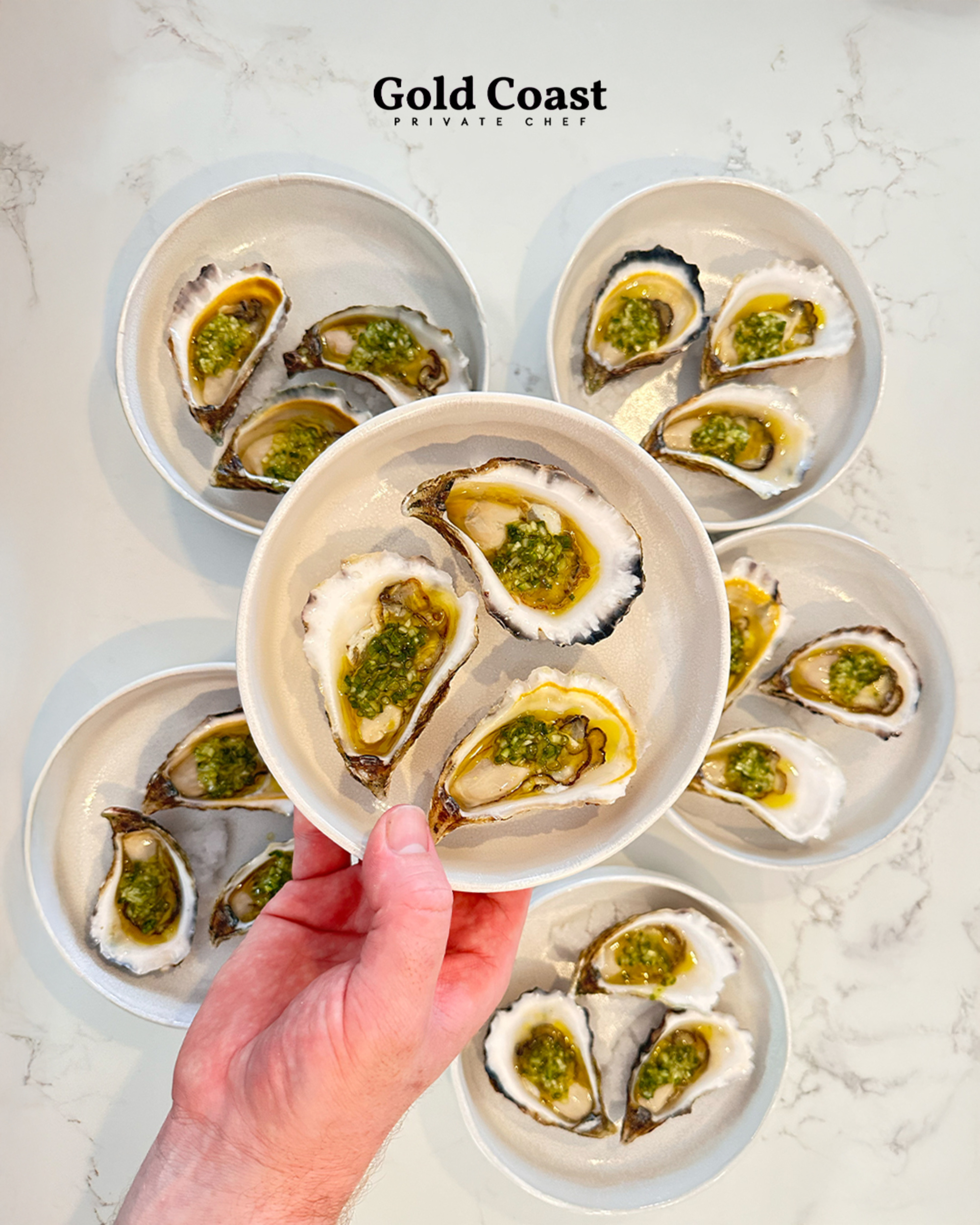 Hand holding a plate of gourmet oysters on the half shell with green garnish, surrounded by matching plates on a marble surface.