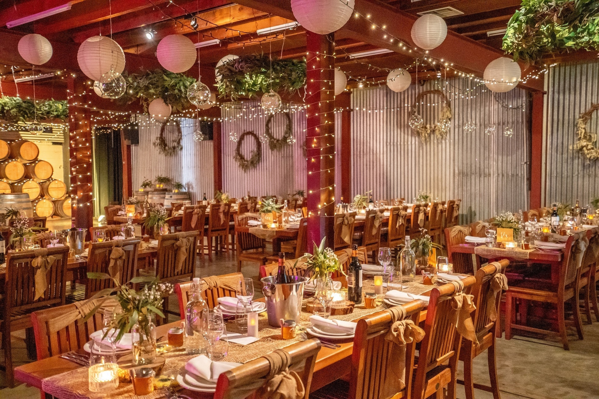 Rustic winery wedding reception with long wooden tables, string lights, greenery, and candlelit decor.