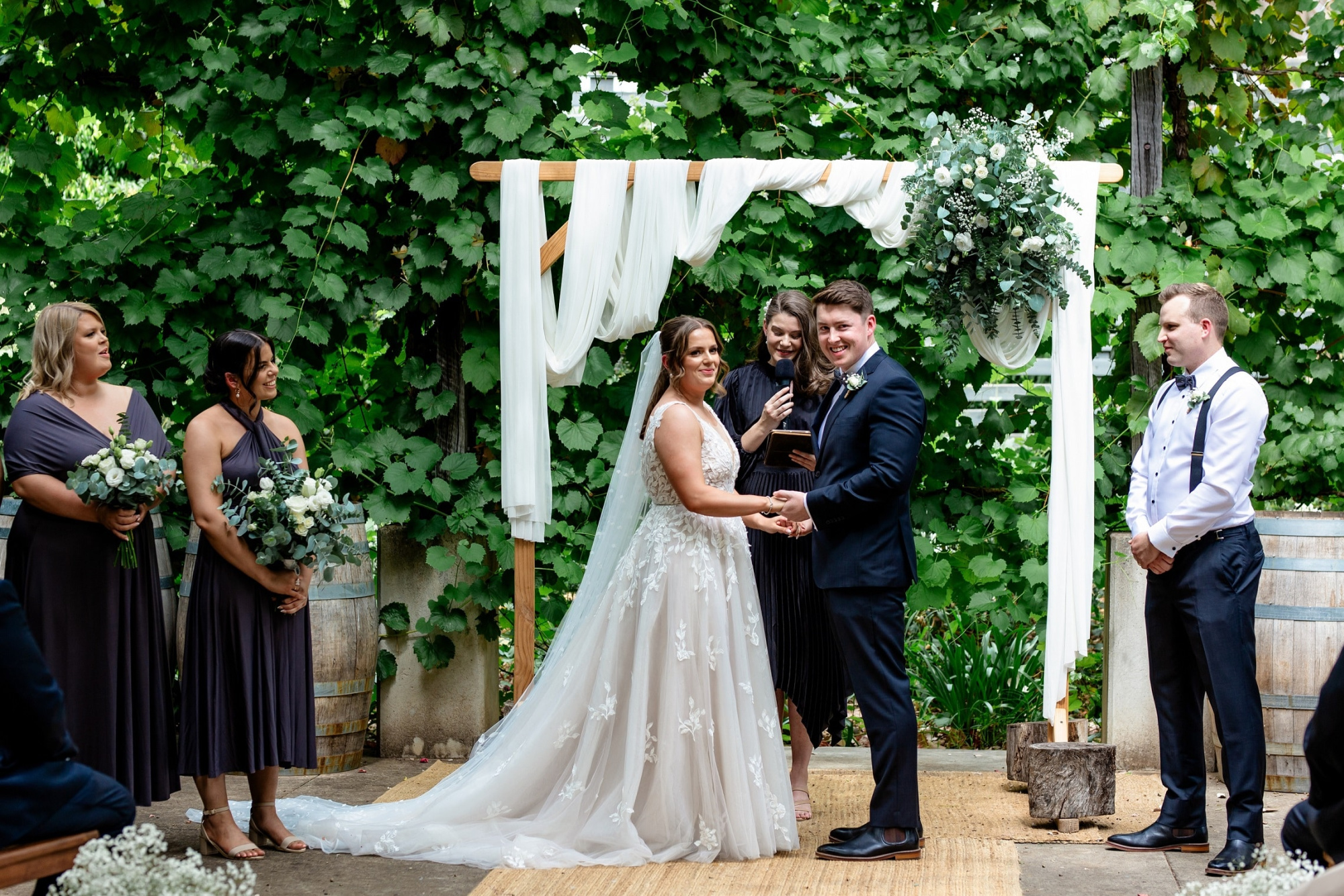 Outdoor wedding ceremony with couple holding hands under a draped wooden arch surrounded by lush greenery and bridal party.