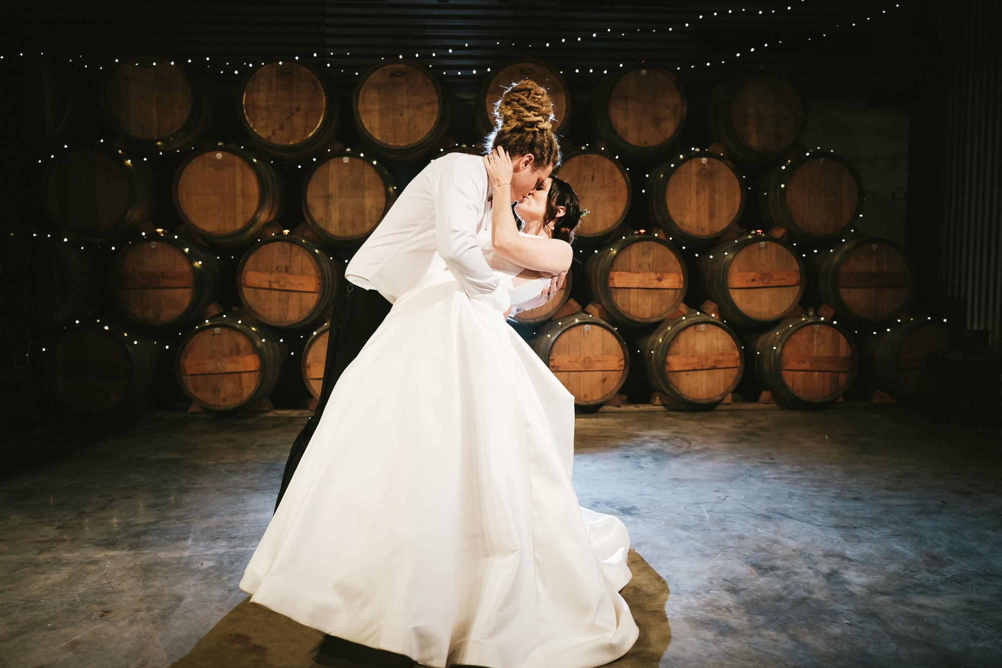 Bride and groom share a romantic kiss during their first dance in a barrel room lit with string lights.