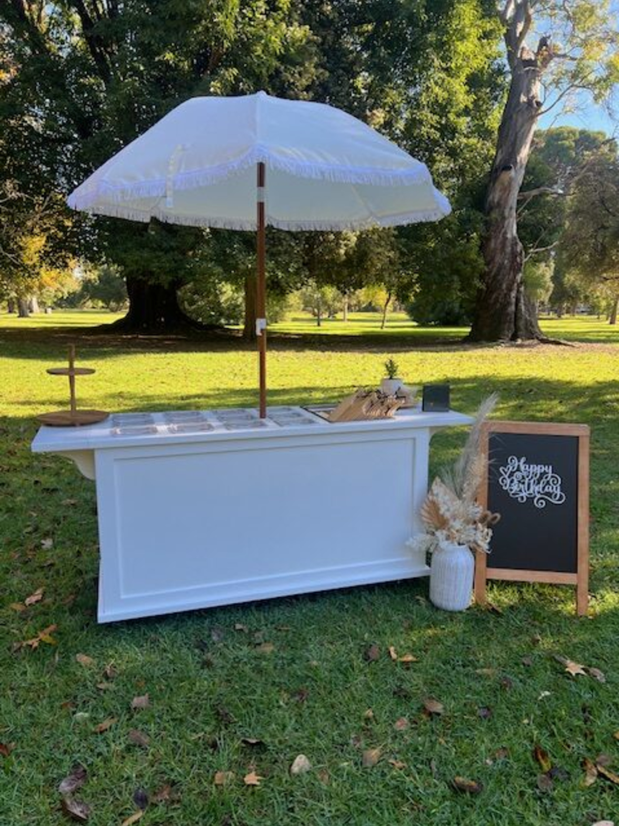 Outdoor white wedding bar with umbrella, chalkboard sign and dried floral arrangement set in a green park.