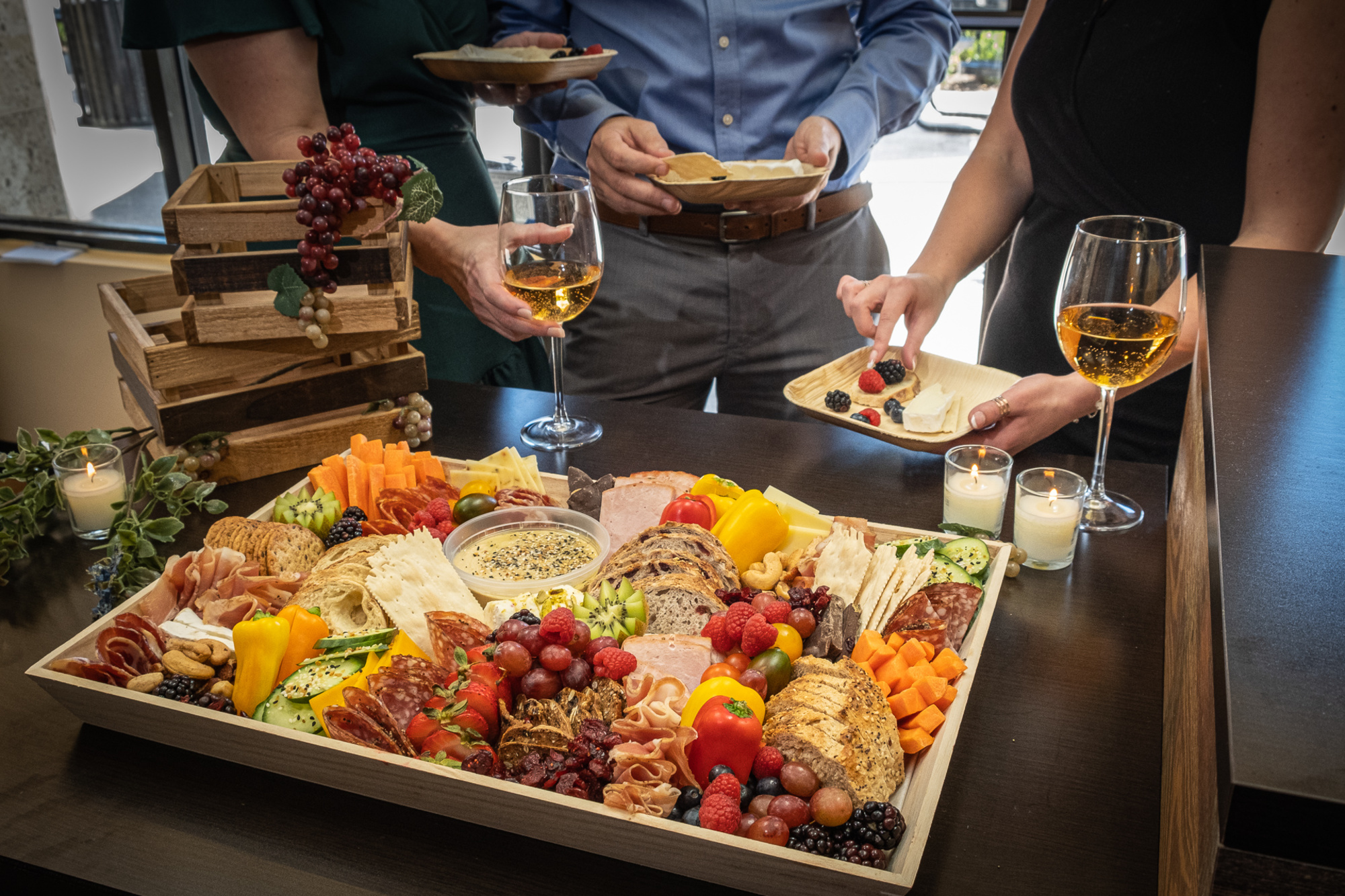 Guests serve themselves from an abundant charcuterie grazing board with wine glasses at a wedding reception.