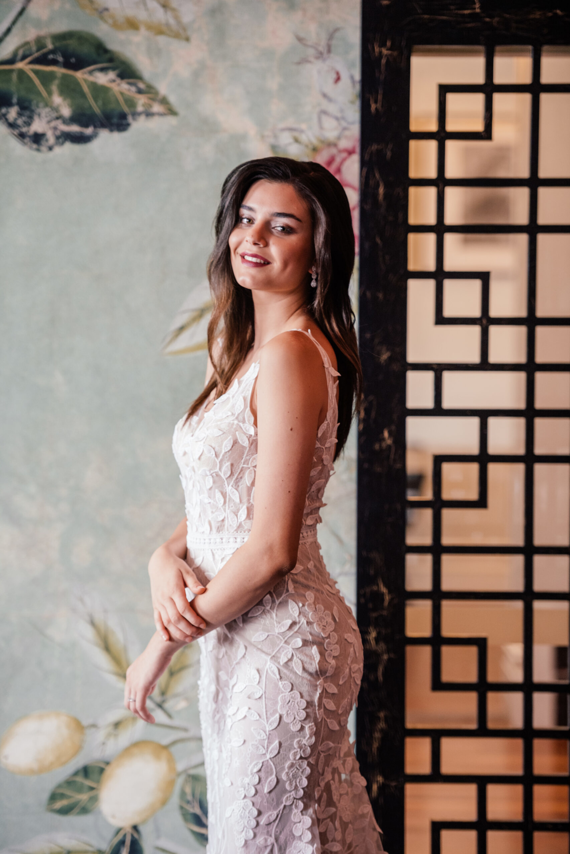 Smiling bride in a fitted lace wedding dress poses indoors against a decorative wall and screen.