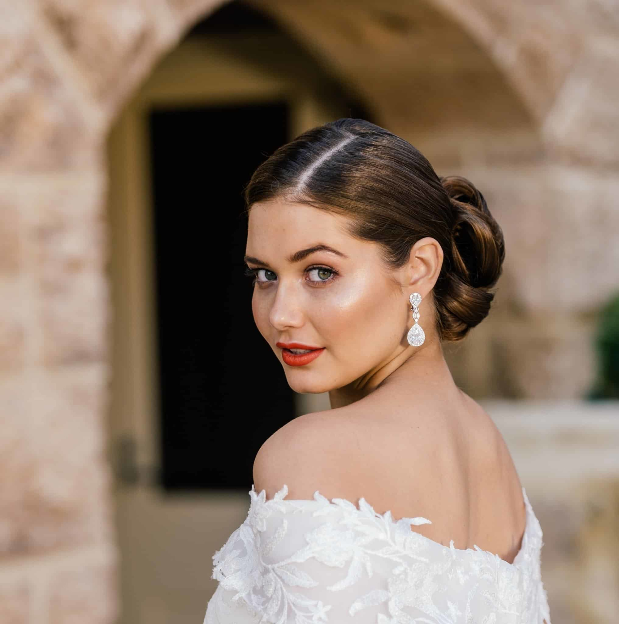 Elegant bride in an off-the-shoulder lace gown with polished updo and statement earrings looking over her shoulder.